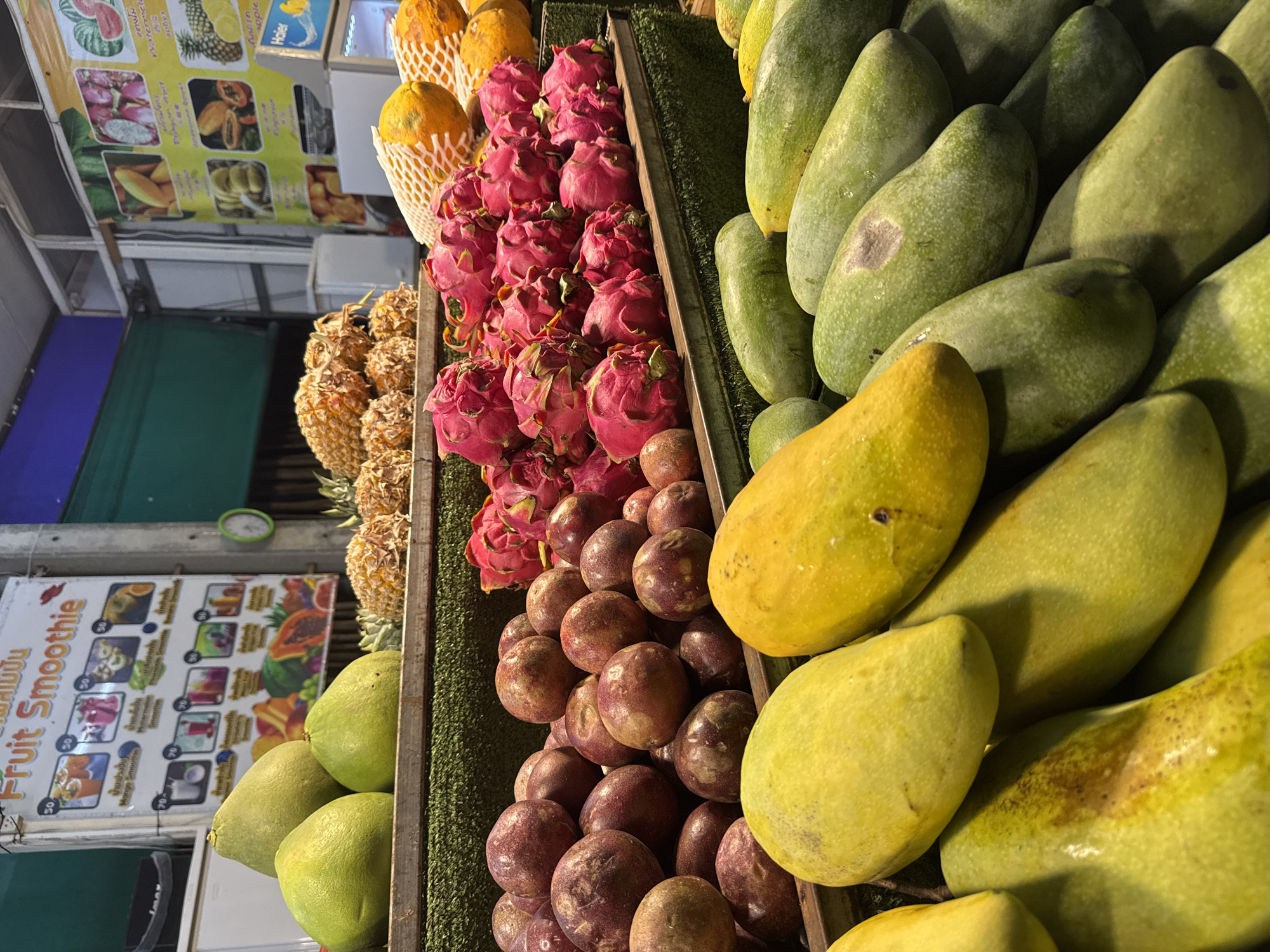 The image depicts a fruit stall in Rawai, Thailand, likely at night given the artificial lighting. The stall features a diverse array of fresh fruits displayed in wooden crates and on tiered shelving. In the foreground, a large selection of green and yellow mangoes are prominently arranged. Behind them, a pile of reddish-purple passion fruits is visible, alongside vibrant pink dragon fruits. Further back, pineapples are stacked, and several papayas, some covered in netting, are also on display. Above the fruit, a sign advertising "Fruit Smoothie" in Thai and English, along with images of various smoothies and fruit ingredients, is mounted on a light-colored structure. Other signs with fruit imagery and pricing are also present. The overall environment appears to be an outdoor market stall.