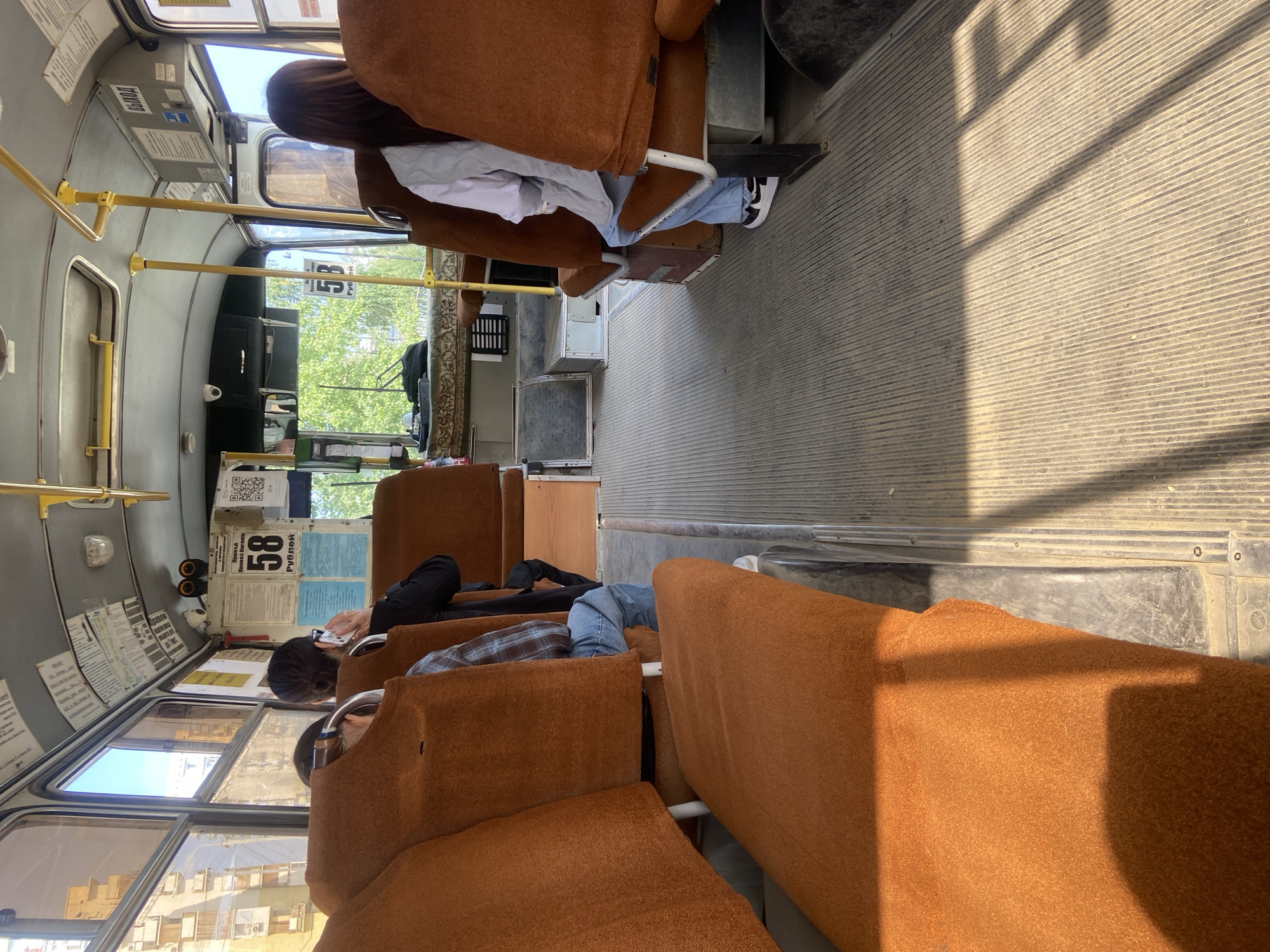 The image shows the interior of a public bus, likely in Yakutsk, Russia, given the Cyrillic text visible on signs. The bus has rows of orange, upholstered seats. In the foreground, the aisle and the floor are visible, with ribbed flooring. Sunlight streams in through the windows, casting shadows on the floor and seats.
Several passengers are seated:
* On the left, a person wearing a blue and plaid shirt and jeans is seated, facing forward.
* Behind them, a person is seated with their back to the camera.
* In the middle of the bus on the right side, a woman wearing a white shirt and blue jeans is seated, facing forward.
* To her right, another woman with long brown hair, wearing a dark jacket and jeans, is seated and talking on a cell phone.
Various signs are attached to the ceiling and walls of the bus. One sign displays the number "58" in large print, followed by "РУБЛЕЙ" (RUBLES) in smaller text. Other signs are in Cyrillic, including one that says "ВЫХОД" (EXIT). A security camera is visible mounted on the ceiling. Outside the windows, trees and buildings can be seen, indicating an urban environment. The overall impression is of a typical scene inside a public bus in Russia during daylight hours.