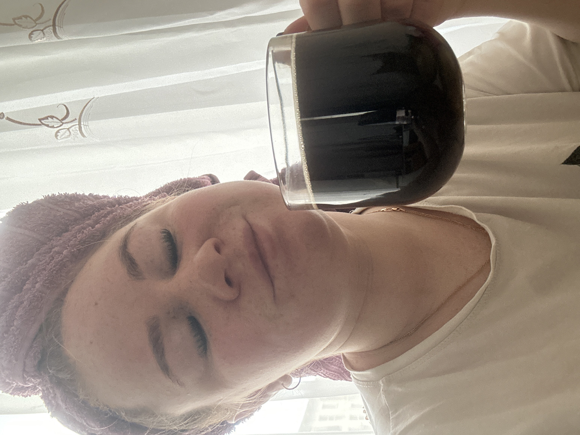 A woman is seen close-up, her face filling much of the frame. She has fair skin, freckles, and her eyes are closed. She is wearing a lavender-colored towel wrapped around her head. She's also wearing a white t-shirt and a thin gold necklace is visible around her neck. Held above her face and slightly tilted is a clear glass mug filled with a dark liquid, likely coffee. In the background, there is a white curtain with a subtle leaf-like pattern. Beyond the curtain, there is a glimpse of a building seen through a window. Based on architectural elements visible in the photo, the location is Chaikovsky, Russia. The interaction is unclear, though the woman is holding the mug of liquid, it is not clear if she is about to drink it.