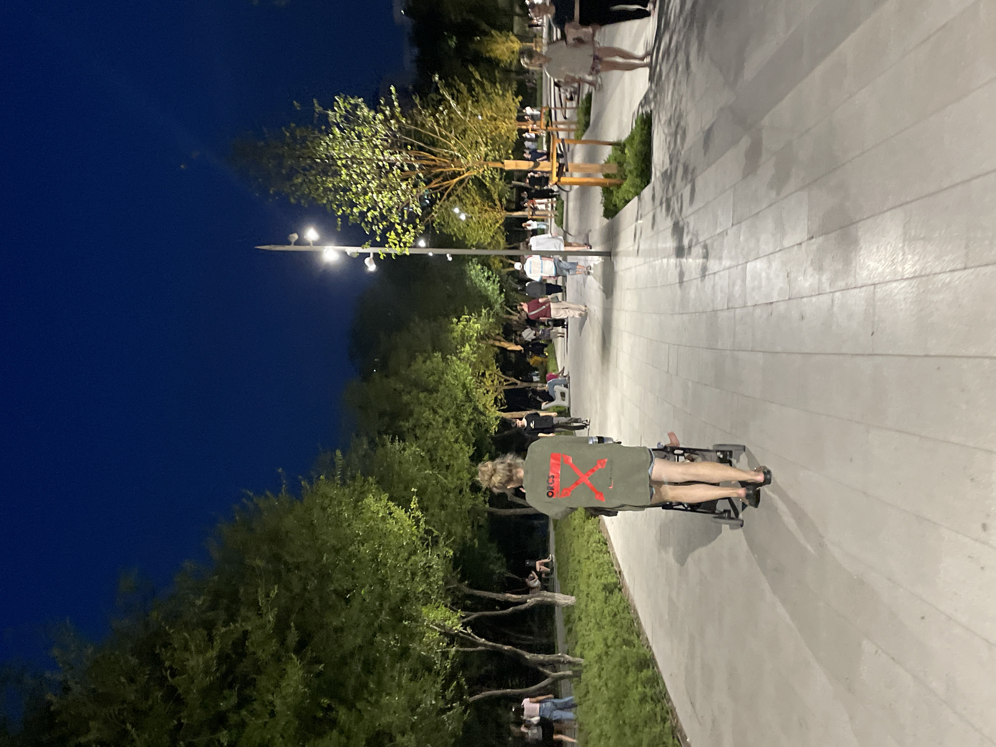 In Almaty, Kazakhstan, a person is seen from behind pushing a stroller down a paved walkway in a park at dusk. The person is wearing a dark olive-colored t-shirt with a large red graphic design on the back and shorts. The walkway is lined with trees, some of which are illuminated by nearby lighting. Other people are walking in the distance along the same path, and some are seated on benches. The sky is a deep blue, indicating the transition from daylight to night.