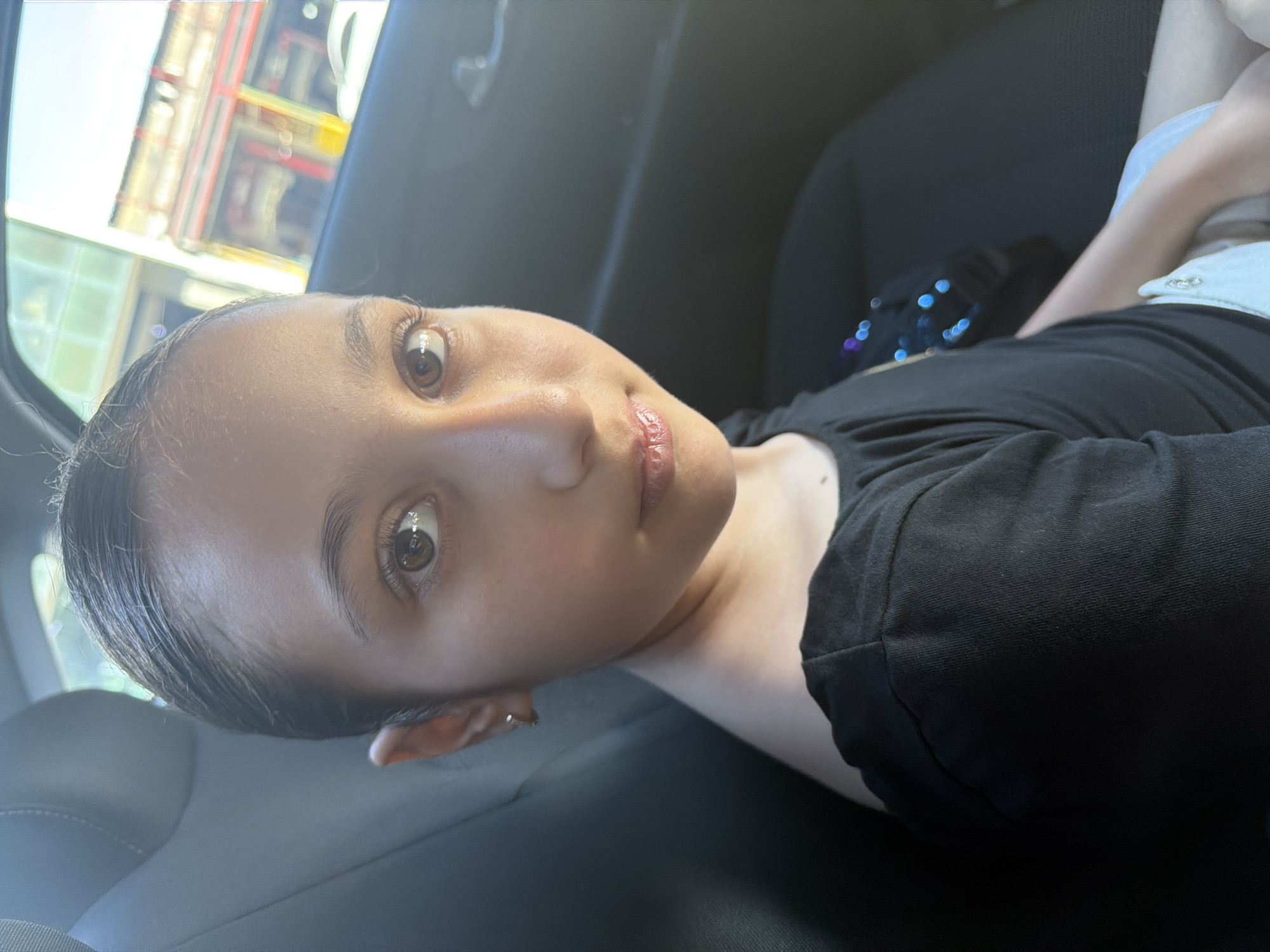 In an indoor setting, possibly a car, a young female of East Asian descent is captured in a close-up shot. She is wearing a black off-the-shoulder shirt. The background includes a car seat and a window, through which a blurry cityscape can be seen, suggesting a location in Yerevan, Armenia. The lighting indicates it's daytime, with sunlight filtering into the scene. The image focuses on the girl's face, highlighting her features.