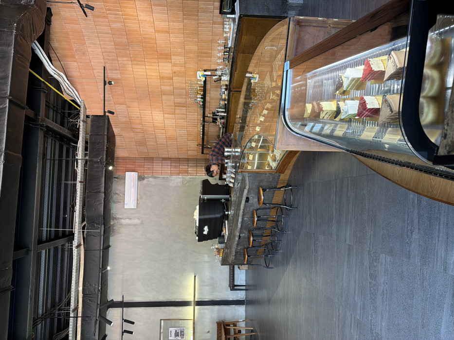 The image depicts the interior of a modern cafe or coffee shop in Kerobokan Kelod, Indonesia. The environment features an industrial aesthetic with an exposed black ceiling structure, including ductwork and track lighting. The left wall is a raw concrete finish, while the right wall is covered in rectangular, light brown terracotta-style tiles. A white air conditioning unit is mounted on the concrete wall. The floor is composed of dark grey, large-format tiles.
A central, curved bar counter with a dark grey, possibly concrete, top is prominent. Four black metal stools with light wooden seats are positioned in front of this bar. Behind the counter, one individual, appearing male, with dark hair and wearing a plaid shirt, is leaning forward, actively engaged with equipment or items on the countertop. A large, black commercial espresso machine is visible on the bar to the left of the individual. Various bottles, glassware, and beverage preparation tools are arranged on shelves behind the bar.
In the foreground and extending towards the mid-ground, two curved glass display cases showcase an assortment of sliced cakes and pastries. On the far left, a wooden table and chairs are partially visible, along with a dark wooden bench against the concrete wall. A framed picture hangs on the concrete wall to the left. The scene is well-lit, suggesting either natural light or ample artificial illumination.