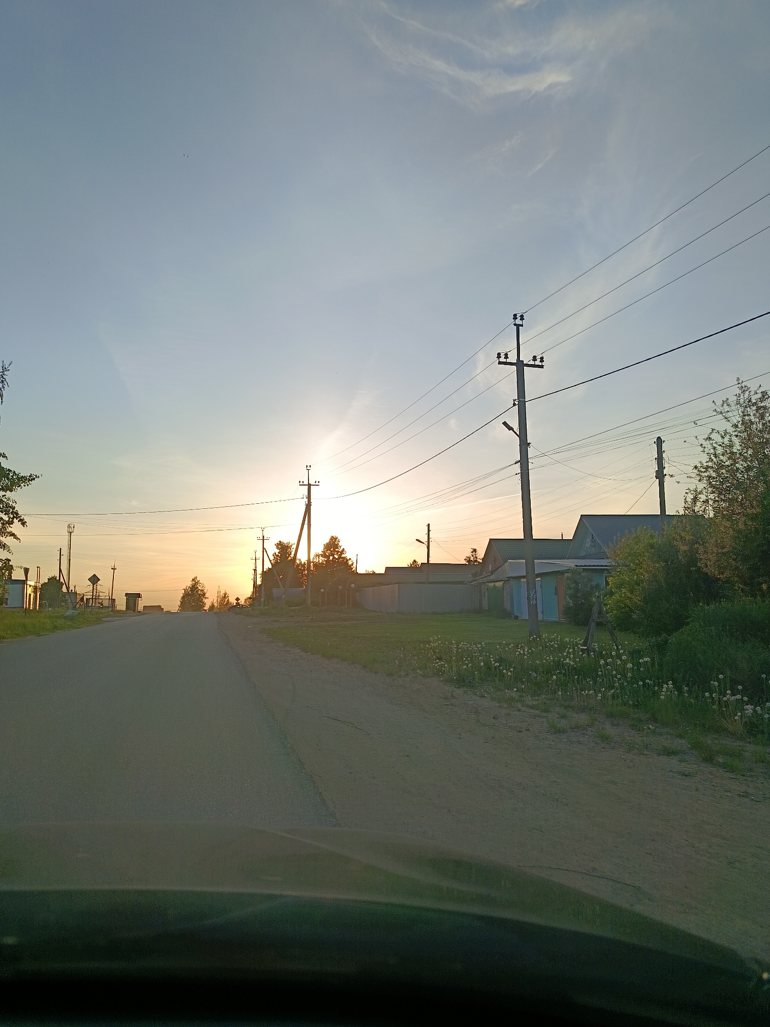 The image depicts a road leading into a village in the vicinity of Chaikovsky, Russia. The location appears to be rural, with houses and power lines flanking the road. The sun is setting, casting a warm, golden glow across the sky and landscape. Dandelions are visible on the side of the road. The photograph appears to have been taken from inside a vehicle.
