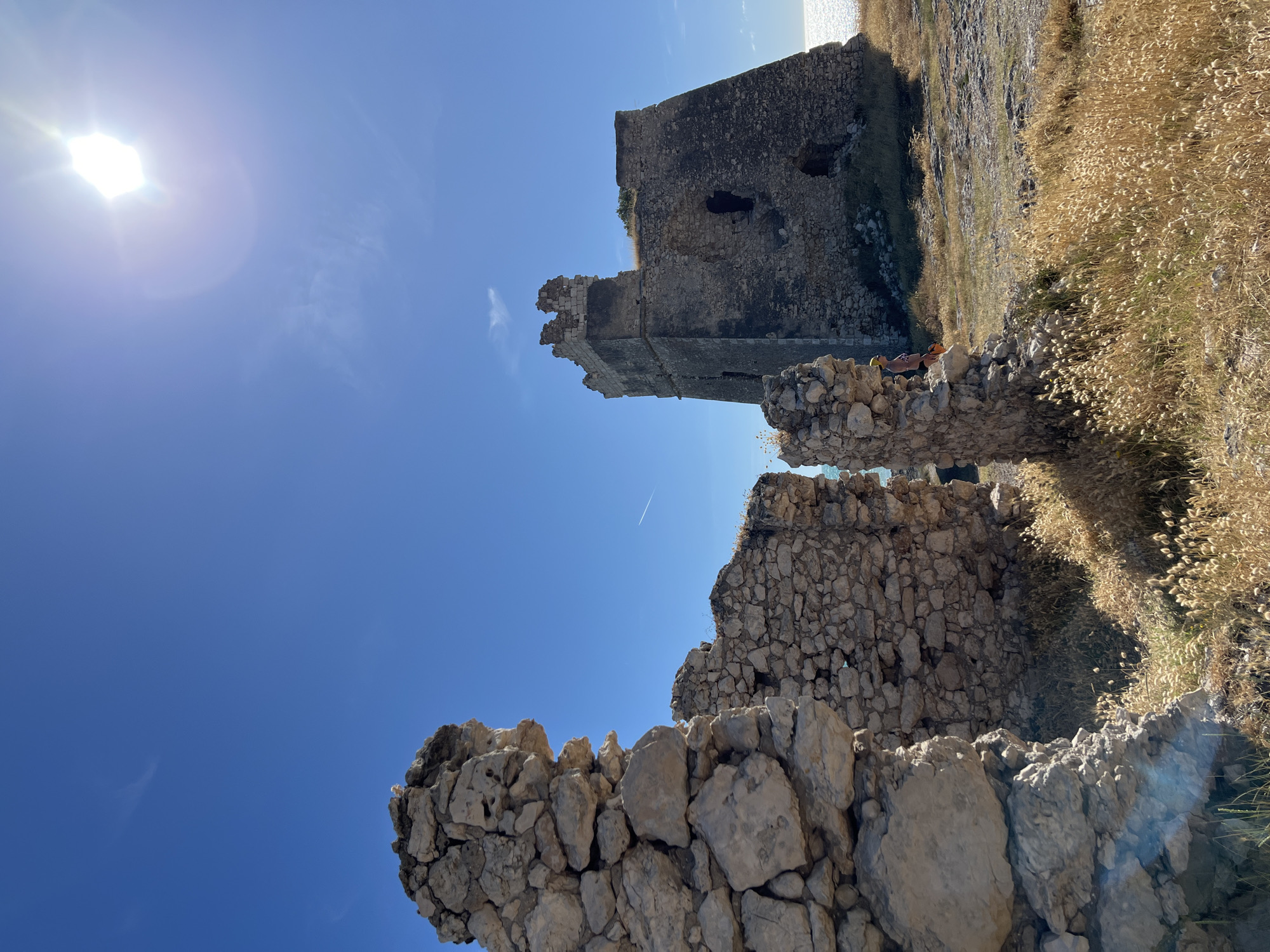 This image depicts the ruins of a stone tower located in Peschici, Italy, overlooking the Adriatic Sea. The tower, partially damaged with crumbled sections at its top, stands on a grassy, rocky terrain. In the foreground, the remains of another stone structure are visible, with large, rough-hewn stones forming walls. Dry, golden grass and sparse vegetation dot the landscape. The sky is a vibrant, clear blue with the sun prominently positioned in the upper right corner, creating a lens flare. A person, partially obscured by the foreground ruins, is visible near the base of the standing tower, wearing a light-colored garment and carrying a bag. The sea glitters in the background under the bright sunlight.