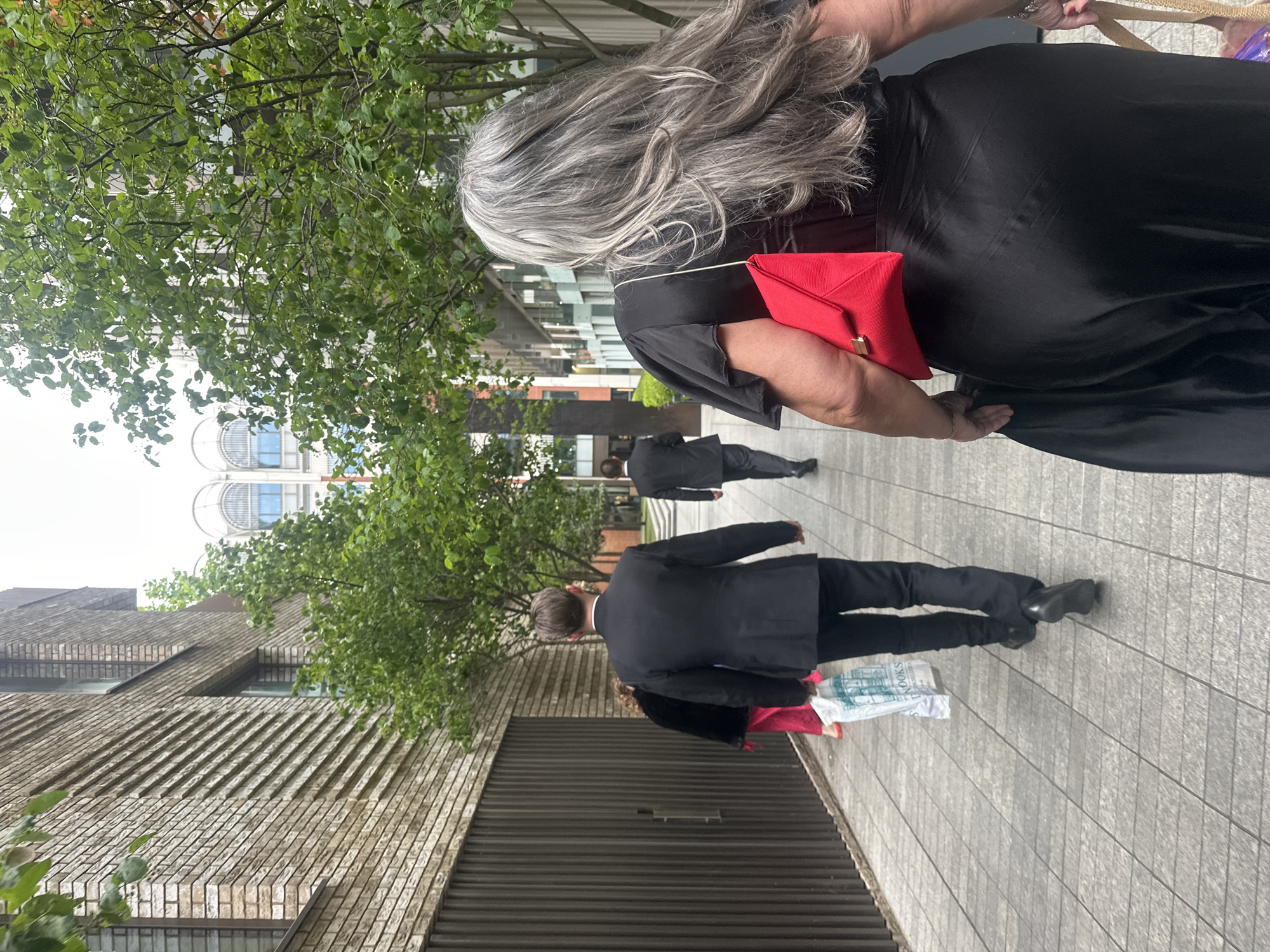 A pedestrian walkway in London, United Kingdom, features a woman with long, gray hair, wearing a black dress and carrying a red clutch. A man in a black suit walks in the opposite direction carrying shopping bags. Another man in a suit walks further away. The environment includes a brick building, a tree, and a glimpse of an upper section of a modern building. The walkway is paved with rectangular stones. The sky is overcast.