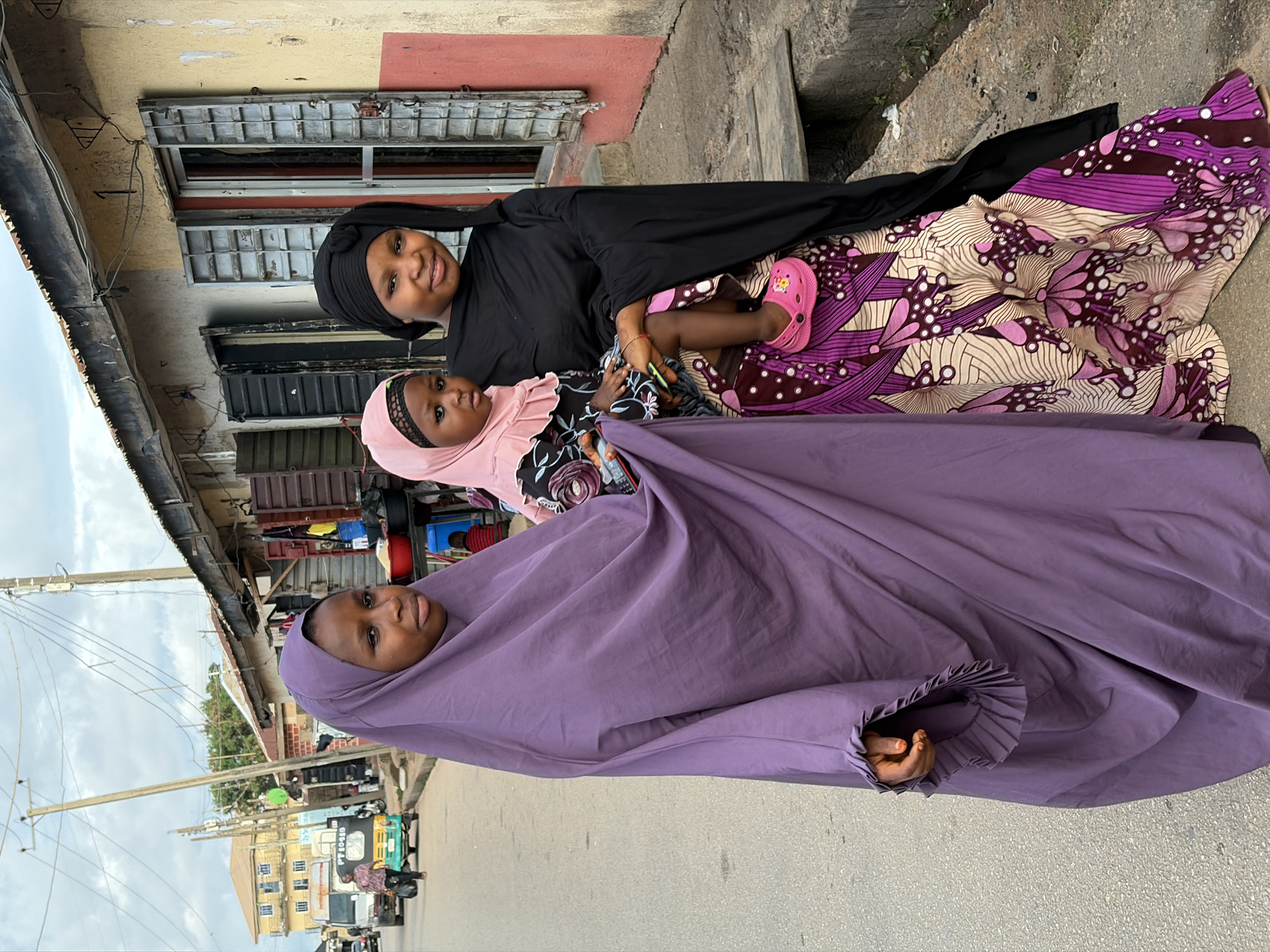 This vertical street-level photograph depicts three individuals—two adult females and one child—standing on a paved road beside a row of buildings in Jos, Nigeria, under an overcast sky.
The adult female on the left, with light-brown skin, wears a full, pleated purple abaya that covers her head and body. Her right hand, adorned with a small ring on the ring finger, is visible near her waist, holding the garment. She looks directly towards the viewer with a neutral expression.
The adult female on the right, also with light-brown skin, wears a black head covering and a black top, paired with a vibrant, floor-length skirt featuring an intricate pattern of large purple, pink, and beige shapes. She carries a small, dark-skinned child on her left hip. She smiles gently towards the viewer. The child, approximately two to three years old, wears a pink head covering, a patterned dress, and bright pink Crocs-style sandals. The child holds a black smartphone in their right hand and looks slightly left with a serious expression.
The immediate background to the right features a weathered, single-story building with faded yellow and red paint, a dark roofline, and metal-shuttered windows. The number "1" is visible above a door. Red plastic bowls and black containers are positioned outside, possibly indicating a street vendor or shop. Further down the street, other similar buildings with dark shutters are visible.
The background to the left reveals a paved street with a grey van and a green and yellow auto-rickshaw (Keke Napep) in the distance. A person walks away from the camera near the auto-rickshaw. Beyond, a multi-story yellow building with blue window frames is visible. Multiple wooden utility poles with a network of overhead electrical wires crisscross the upper frame against the grey, cloudy sky.