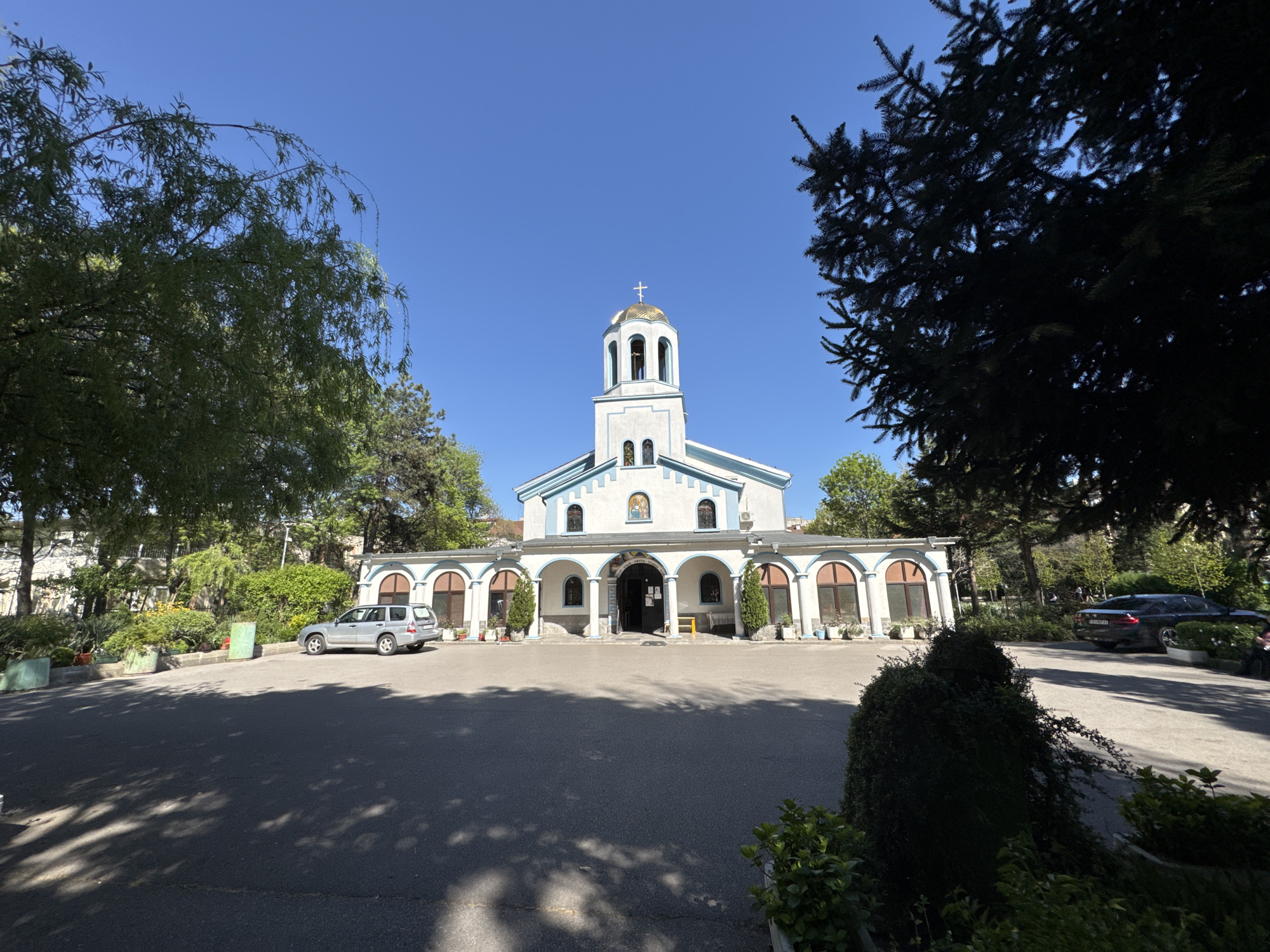 The image shows the Church of the Holy Mother of God in Sofia, Bulgaria, under a clear blue sky. The church is white with blue trim, featuring a dome topped with a golden cross and an arched portico supported by columns. A parking area is in front of the church. A silver SUV is parked in the parking area to the left of the image. A black car is parked at the right edge of the image. Green trees and shrubbery surround the perimeter of the area. Shadow patterns from the trees are visible on the parking area.
