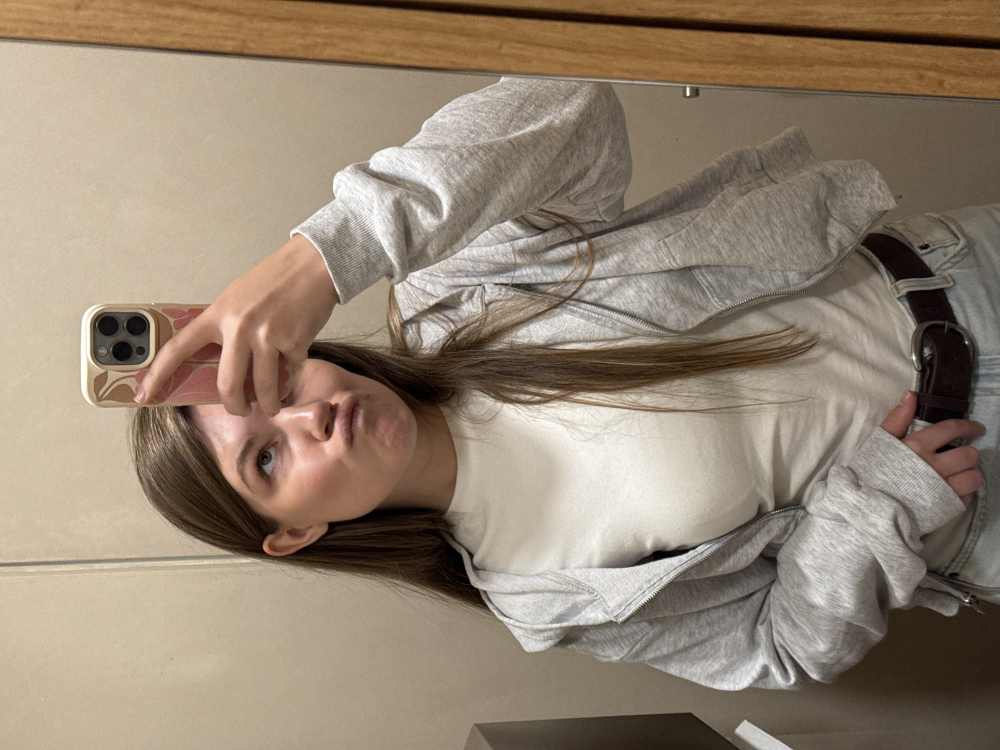 A woman is taking a selfie in a mirror. She is wearing a white t-shirt, a grey zip-up hoodie, light blue jeans, and a brown belt. She is holding a phone with a patterned case up to her face. The location cannot be confirmed as Vracar Urban Municipality, Serbia from the provided image.