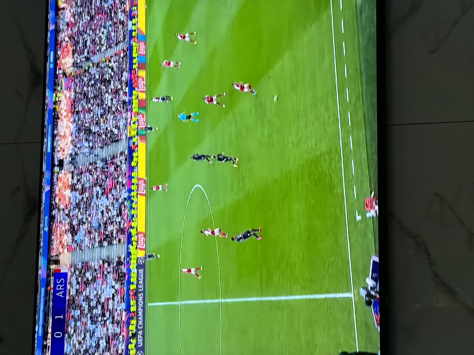 The image shows a television screen displaying a soccer game, specifically a UEFA Champions League match. The score is 0-1, with "ARS" displayed, suggesting Arsenal is involved. The view is from above, showing the green soccer field, players in various positions, and the crowd in the stands. Advertisements line the side of the field, including "Lays". The image's setting is indoors, showing a glimpse of a marble or tiled wall next to the TV screen. There's no indication the location is in the city of Asenovgrad, Bulgaria.