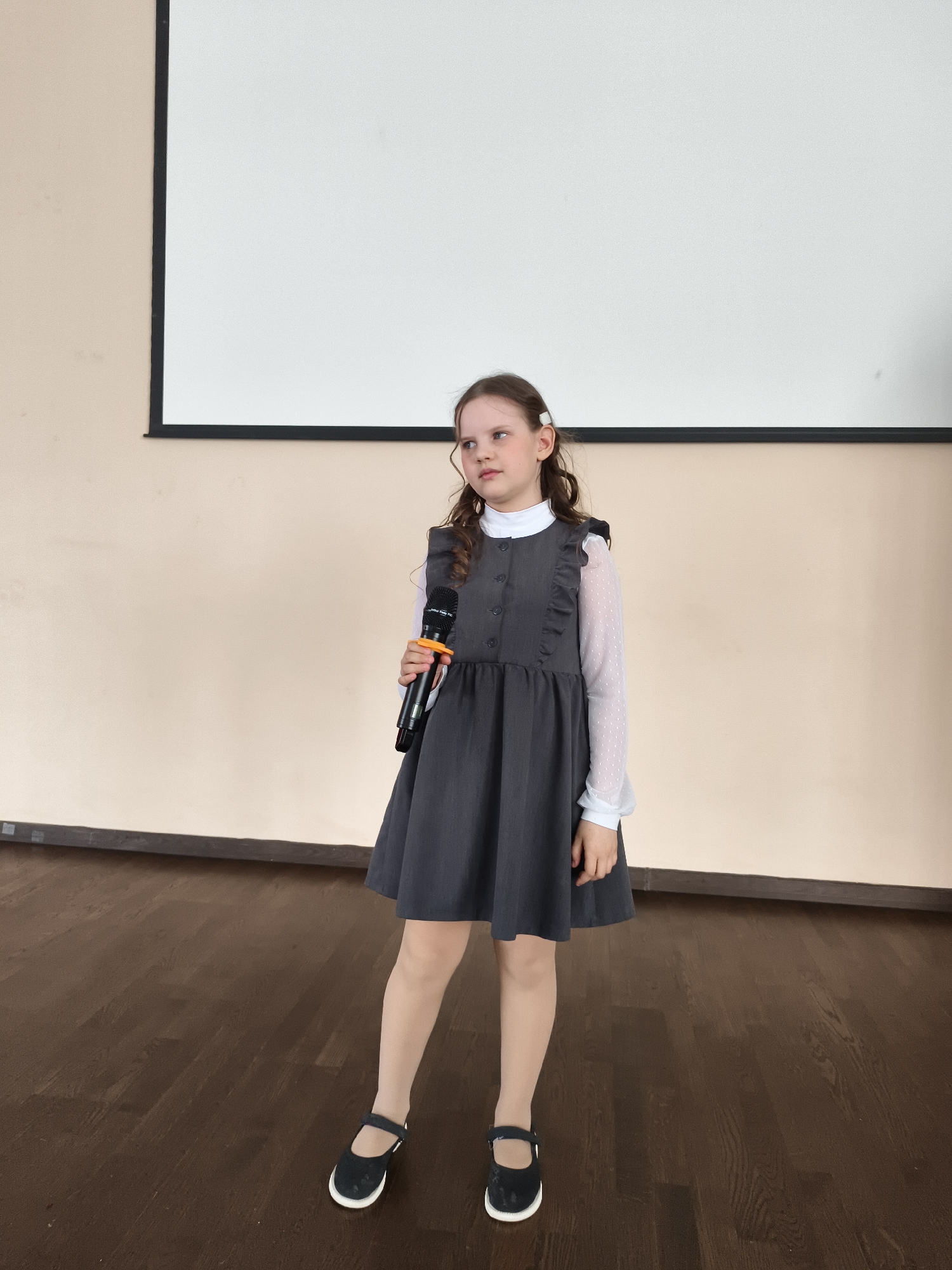 A young girl stands on a dark brown wooden floor, holding a black microphone with an orange band in her right hand. She has long, wavy brown hair, secured with a white clip on the right side of her head, and is looking slightly towards her left (viewer's right) with a thoughtful expression. She is wearing a grey pinafore dress with button details and ruffles on the shoulders, layered over a white long-sleeved top with a high collar and sheer, subtly textured sleeves. Light-colored tights and black Mary Jane-style shoes with white soles complete her outfit. Behind her is a light beige wall with a dark brown trim along the bottom. A large, rectangular white projection screen, framed in black, is mounted on the wall behind her, appearing blank. The indoor setting, featuring a projector screen and a clear standing area, suggests a school classroom, assembly hall, or a multi-purpose room where presentations or performances might occur. She appears poised to speak or perform.