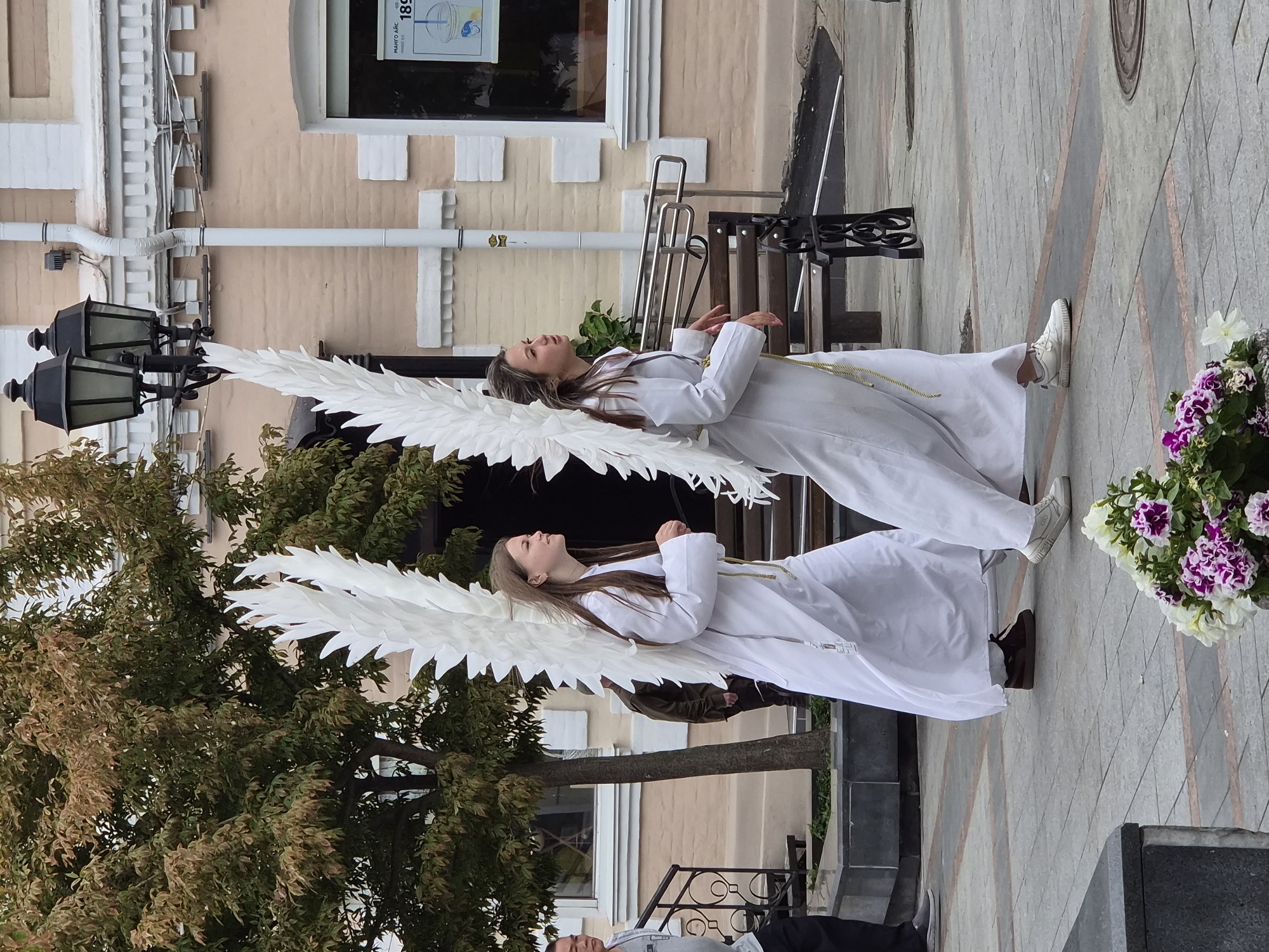 Two young women dressed as angels, wearing long white robes and large white feathered wings, are walking across a paved plaza. The woman on the left has long brown hair and is looking to her right. The woman on the right has lighter brown hair and is looking to her left. They are walking from left to right in the frame. Behind them, a building with a pale pink facade and white trim is visible. The building has windows, and a streetlamp with two black lamps is attached to the side. A tree with green and brown leaves is to the left of the women. In the foreground, a planter with purple and white flowers is visible. The scene appears to be a public outdoor space, possibly a street or square in Vladivostok, Russia.