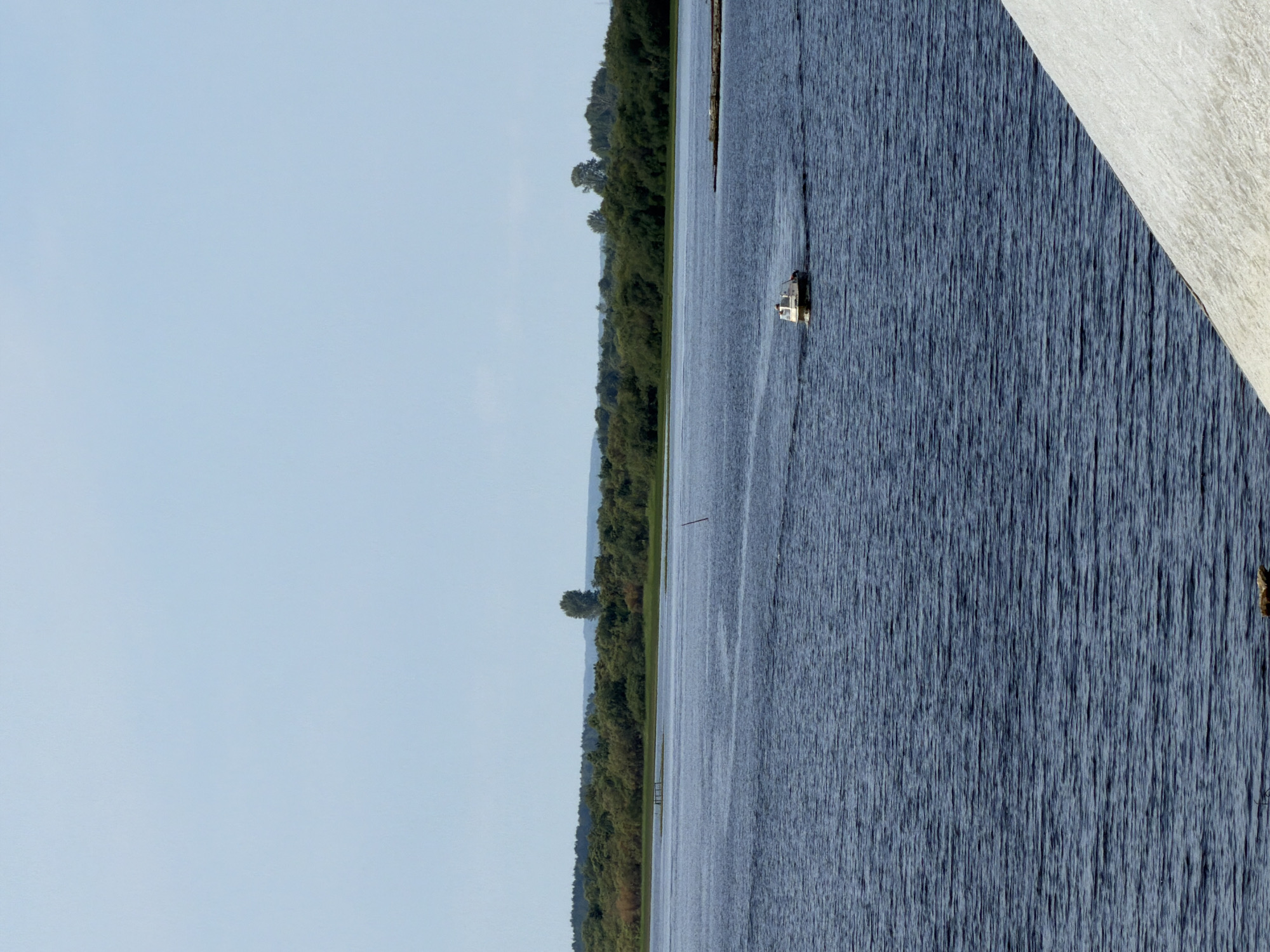 A motorboat navigates a wide, dark blue body of water under a clear, pale blue sky. The water's surface is rippled, suggesting a gentle breeze. In the distance, a dense, green treeline forms the horizon, dotted with a few taller trees. A small, light-colored structure, possibly a pier or breakwater, juts out from the right side of the frame. In the foreground, the edge of a large, light-colored mound, possibly made of salt or snow, slopes upwards from the bottom right corner of the image. The image appears to be taken from a slightly elevated perspective. This scene is the Kama River in Solikamsk, Russia.