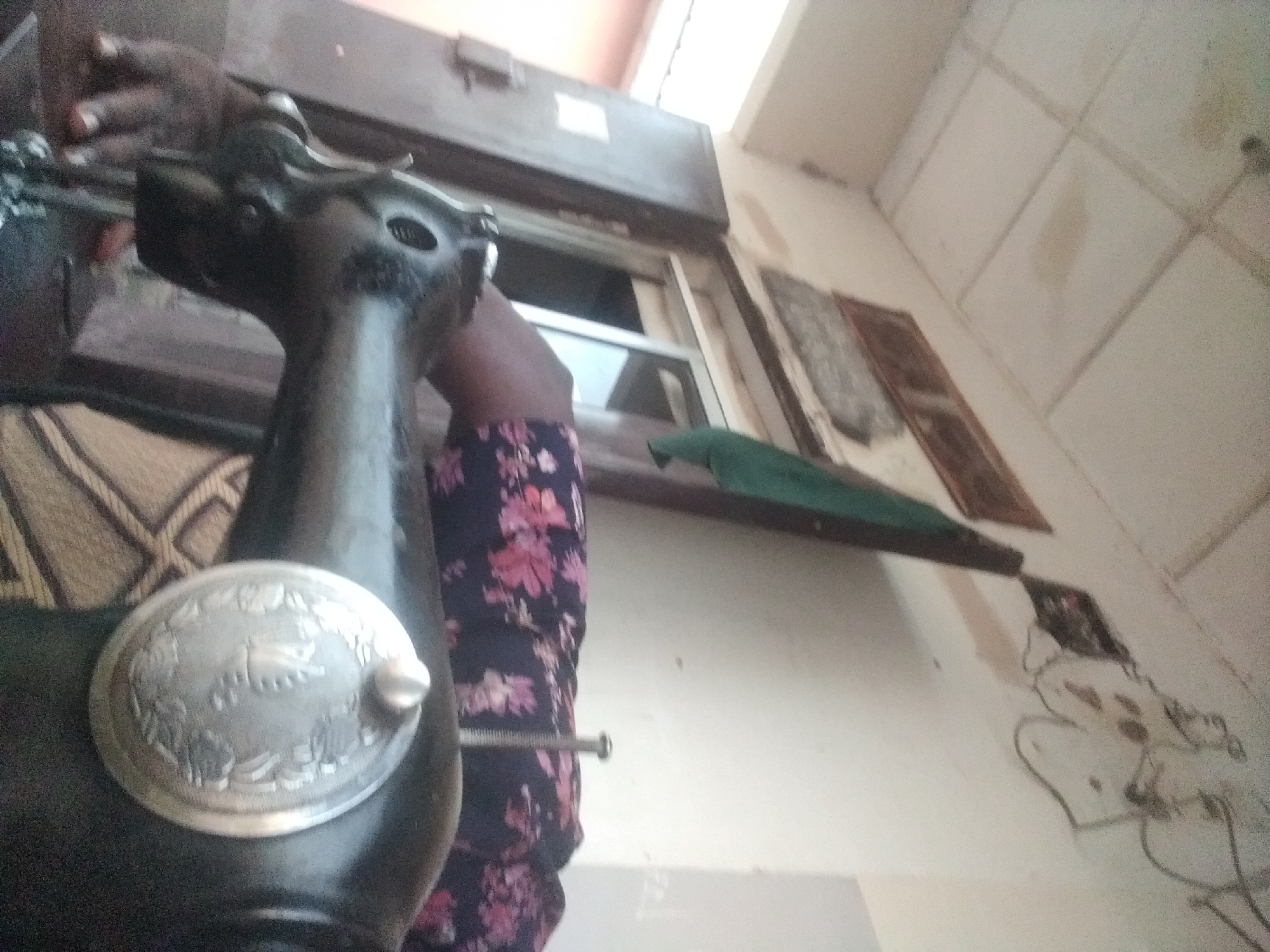 The image captures a close-up view of a vintage sewing machine in an indoor setting. The sewing machine, predominantly black, is positioned in the foreground. A person's hands are visible operating the machine, with one hand on the fabric and the other near the base. The fabric on the machine has a dark background with a floral pattern in shades of pink and red. In the background, a wooden door and a window with a white frame are discernible. The walls appear to be painted a light color, and there is a tiled ceiling above. A green fabric is draped over a pole or support structure in the upper right portion of the image. The overall environment suggests a domestic or workshop setting. The context provided indicates this scene is located in Jalingo, Nigeria.