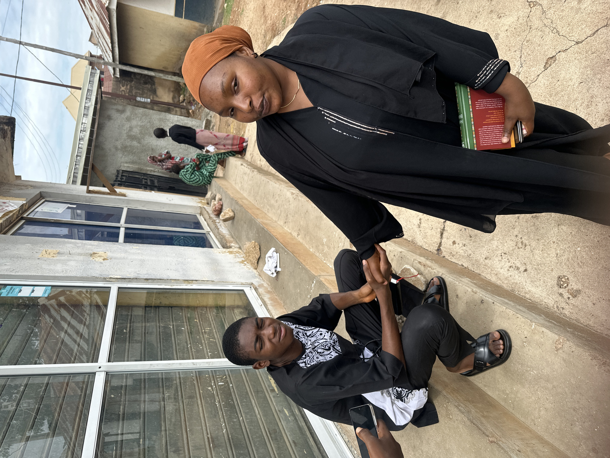 An outdoor, eye-level full shot captures a street scene in the city of Unknown, Unknown. In the foreground, a dark-skinned woman stands on the right, facing slightly left and looking towards the camera. She wears an orange-brown head covering and a black abaya with subtle silver embellishments on the front and sleeve cuffs, holding a stack of books (green, red, and yellow) in her left arm. Her right hand clasps the right hand of a dark-skinned young man seated on a concrete ledge to her left. The young man, wearing a black tunic with white patterns and black trousers, has a pained expression with eyes closed. He wears black open-toed sandals. An unseen hand on the far left holds a black smartphone near his left hand.
The background on the left features a weathered light grey building with large aluminum-framed glass windows covered by closed, distressed grey metal roll-down shutters. A small white "PAY WITH TRANSFER" sticker with a QR code is visible on one window. The building's wall shows crumbling plaster. On the concrete ledge to the left of the young man, several small rocks are present; a crumpled white cloth lies to his right. In the midground, three women are partially visible: one in a green patterned outfit seated on the steps, another standing behind her in a light pink skirt and dark top, and a third with a patterned head covering. Beyond the immediate building, other simple structures with corrugated metal roofs are seen under an overcast sky, with utility poles and wires visible. The ground is worn concrete pavement.