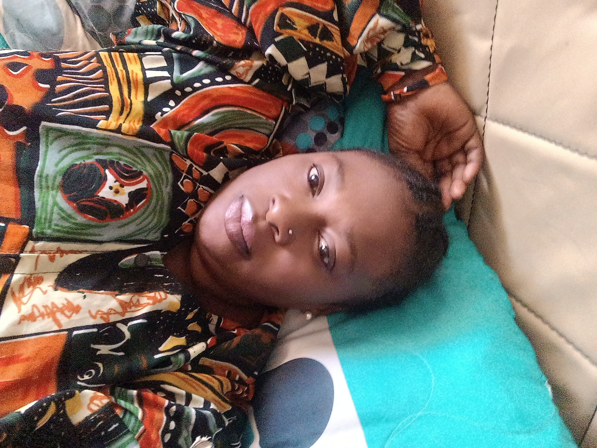 A young woman lies on a patterned fabric surface, her head resting on a teal-colored pillow. She is wearing a brightly colored, patterned top. Her left hand is visible near her head. The location is specified as Jos, Nigeria. The background shows a beige quilted surface.