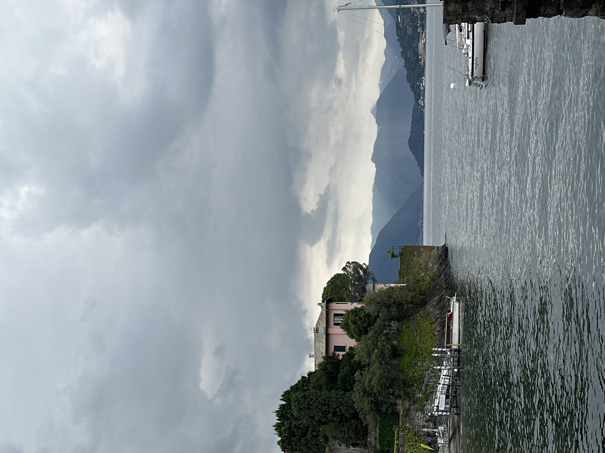A grey, overcast sky looms over a tranquil body of water, likely Lake Como, in Varenna, Italy. On the left side of the image, a pink villa is nestled amongst lush green trees on a rocky outcrop overlooking the water. A wooden dock extends into the water, with several small boats moored to it. The water itself is calm, with gentle ripples disturbing its surface. In the distance, hazy blue mountains form a dramatic backdrop, their peaks softened by the atmospheric conditions. On the right side of the image, a white sailboat with a tall mast is docked, and a stone wall in the foreground suggests a waterfront promenade or structure. The overall impression is one of serene, subdued beauty.