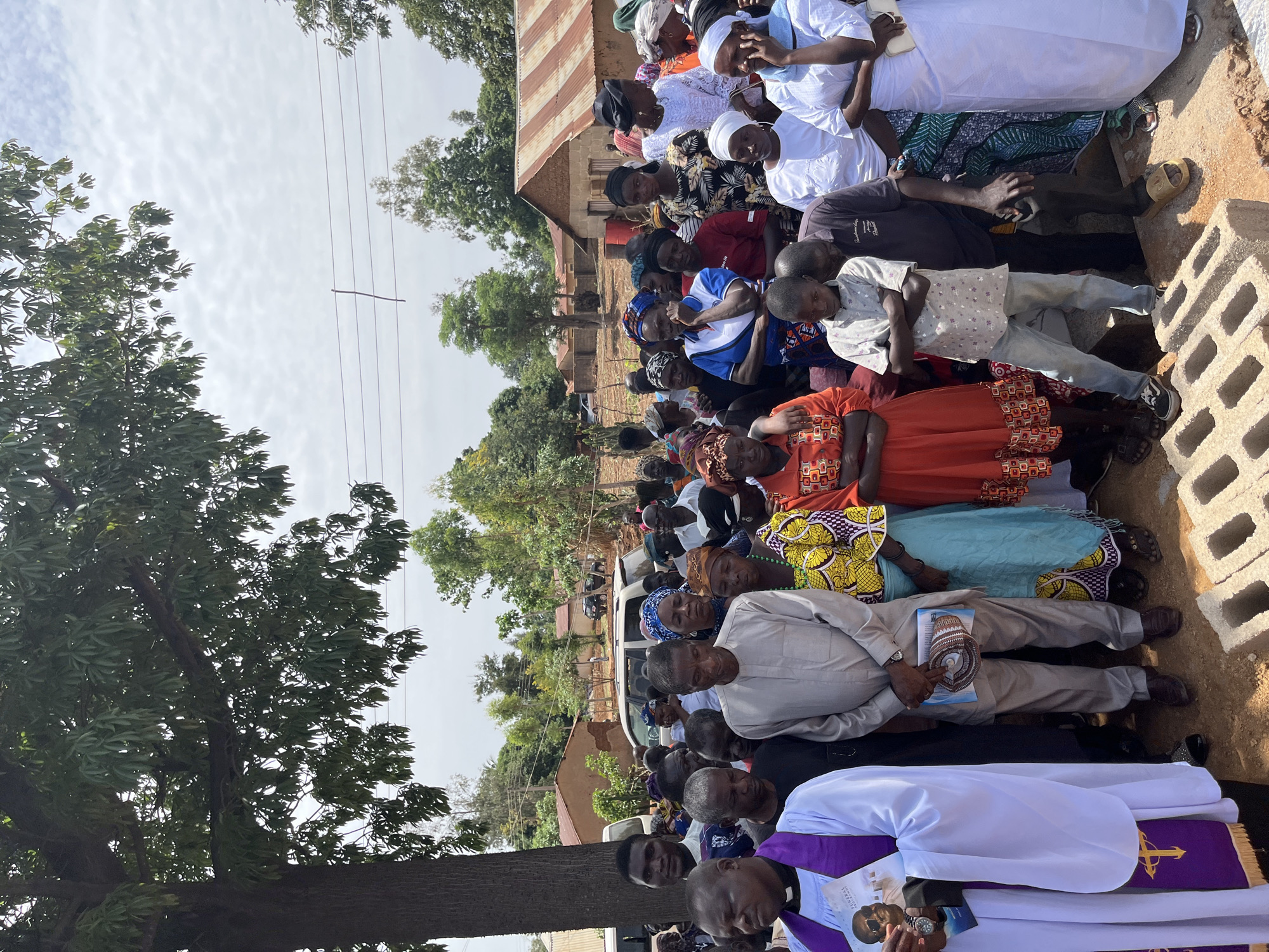 The image depicts a gathering of people in Dogon Dinia, Nigeria. A large group of people of varying ages are assembled outdoors, many of whom are seated. The people are dressed in a mix of traditional African clothing, casual attire, and religious garments. A man wearing a white and purple clerical collar appears to be a religious leader, holding what looks like printed materials. The gathering is taking place near buildings with corrugated metal roofs, and a parked white van is visible in the background. A large tree stands on the left side of the frame. The weather appears to be clear and sunny.