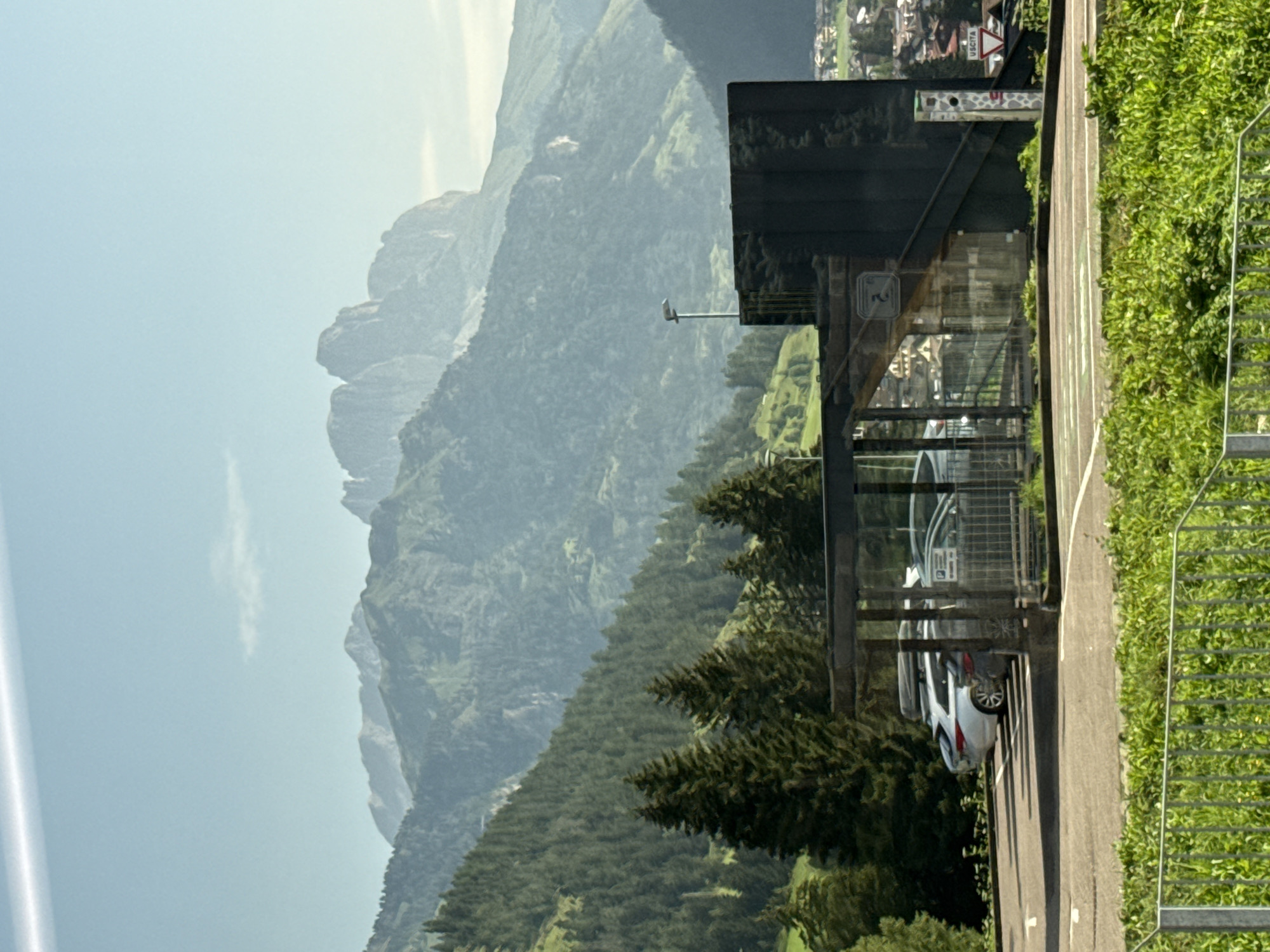 In San Giovanni di Fassa, Italy, a covered parking area is situated in front of a backdrop of lush green mountains and a clear sky. The parking area features a modern, dark-colored structure with a sloped roof and glass enclosures. Several cars are parked, including a white SUV with a roof rack and a silver sedan. A metal fence with green foliage in the foreground suggests the edge of the parking lot. In the background, the rugged peaks of the Dolomites rise majestically, partially obscured by a light haze. A signpost with the word "USCITA" (exit in Italian) is visible near the parking area. The scene captures a typical day in a mountainous region, where natural beauty meets infrastructure.