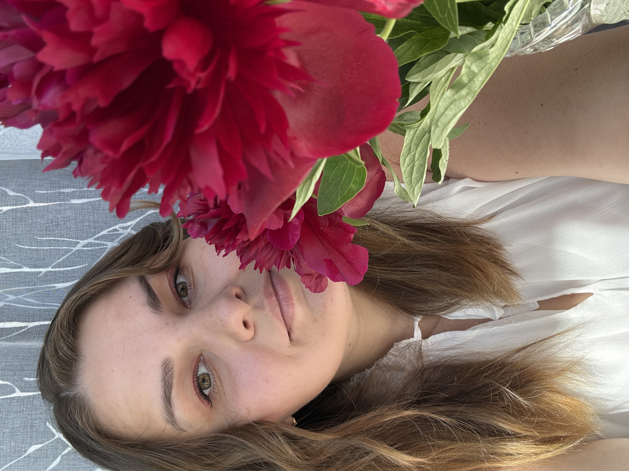 A young woman with light brown hair and green eyes lies on a grey patterned surface. She wears a white shirt. A large red peony is held above her head, partially obscuring her face. The scene is brightly lit. There is no indication of the location as Chaikovsky, Russia within the image.