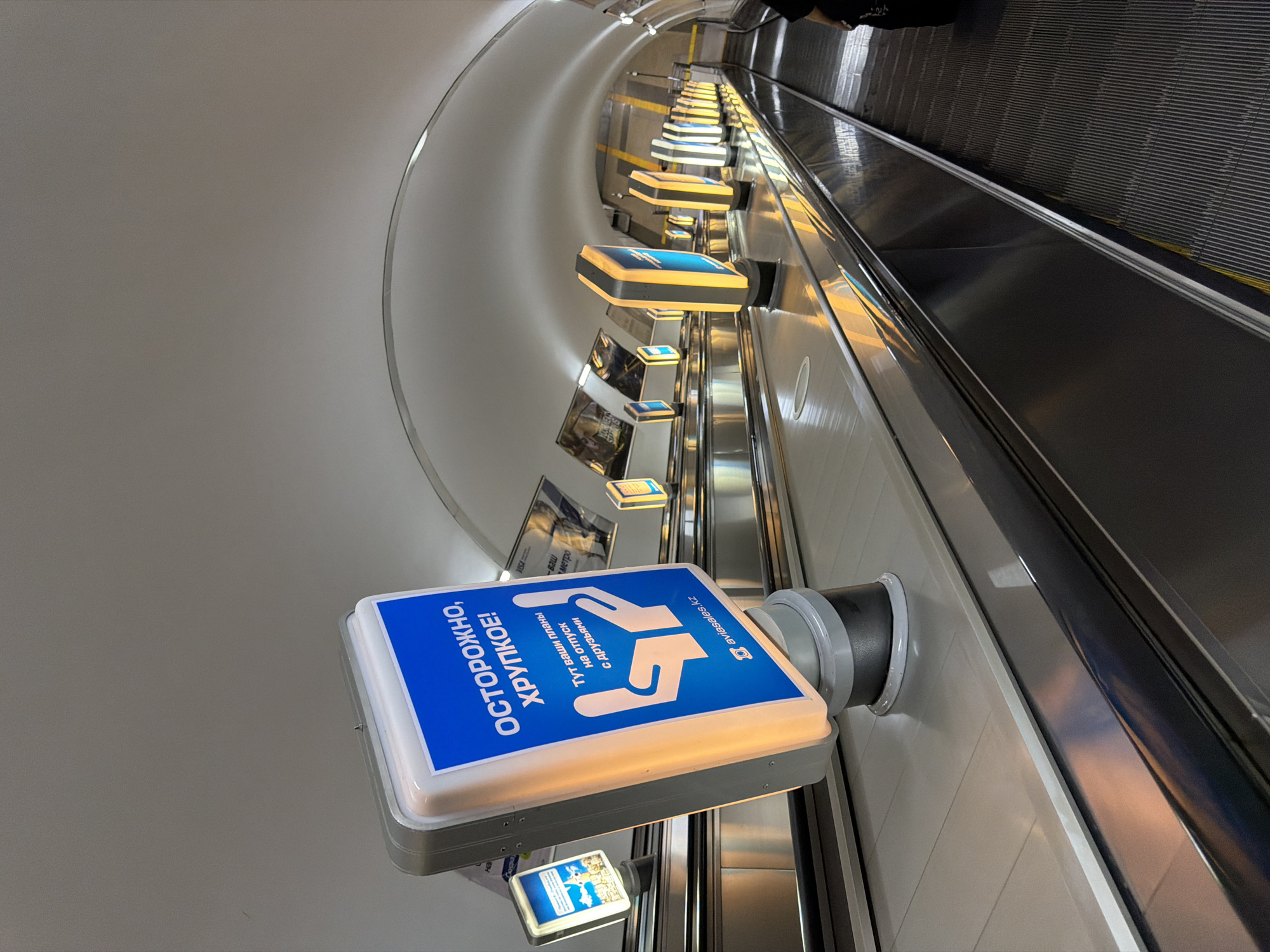 An upward-sloping escalator descends into a tunnel-like environment, illuminated by a series of rectangular, backlit signs. The closest sign, prominently displayed, features a blue background with white text and graphics. The text reads "ОСТОРОЖНО, ХРУПКОЕ!" which translates to "CAUTION, FRAGILE!". Below this, smaller text says "Тут ваши планы на отпуск с друзьями" ("Here are your plans for a vacation with friends"). The graphic depicts two cupped hands, suggesting care and fragility. A website address, "aviasales.kz", is visible at the bottom of this sign. Other similar signs, also with blue and yellow illumination, are spaced along the escalator's sides, receding into the distance. The escalator treads are dark and ribbed, contrasting with the smooth, reflective metal of the side panels. The ceiling of the tunnel is a smooth, curved, white surface. In the far distance, the yellow lines marking the edge of the escalator are visible. A person's legs, wearing dark clothing, are visible at the bottom right of the frame, indicating movement down the escalator. The overall scene suggests a public transport hub or a commercial area, specifically Almaty, Kazakhstan, given the ".kz" domain on the sign.