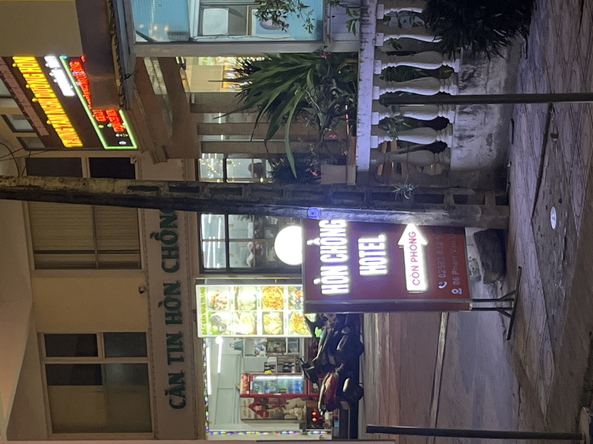 The image depicts a street view at night in Bac Nha Trang Ward, Vietnam. In the foreground, a maroon sign with white lettering announces "HON CHONG HOTEL". An arrow on the sign points to the right, with the Vietnamese text "CON PHONG" (meaning "room available") illuminated in white. Below this, a phone number "02582 832 5" and an address "06 Pham Van D" are visible. A metal pole stands to the left of the sign.
Behind the sign, a row of white balusters borders a raised concrete area with plants. To the left of this, several motorcycles are parked. Further back, the facade of a building features large windows and a sign with green and yellow neon lettering that appears to be advertising "TRUNG TAM NGHI DUONG HON". A red refrigerator unit is visible through a storefront window. The overall scene is illuminated by the neon signs and streetlights.