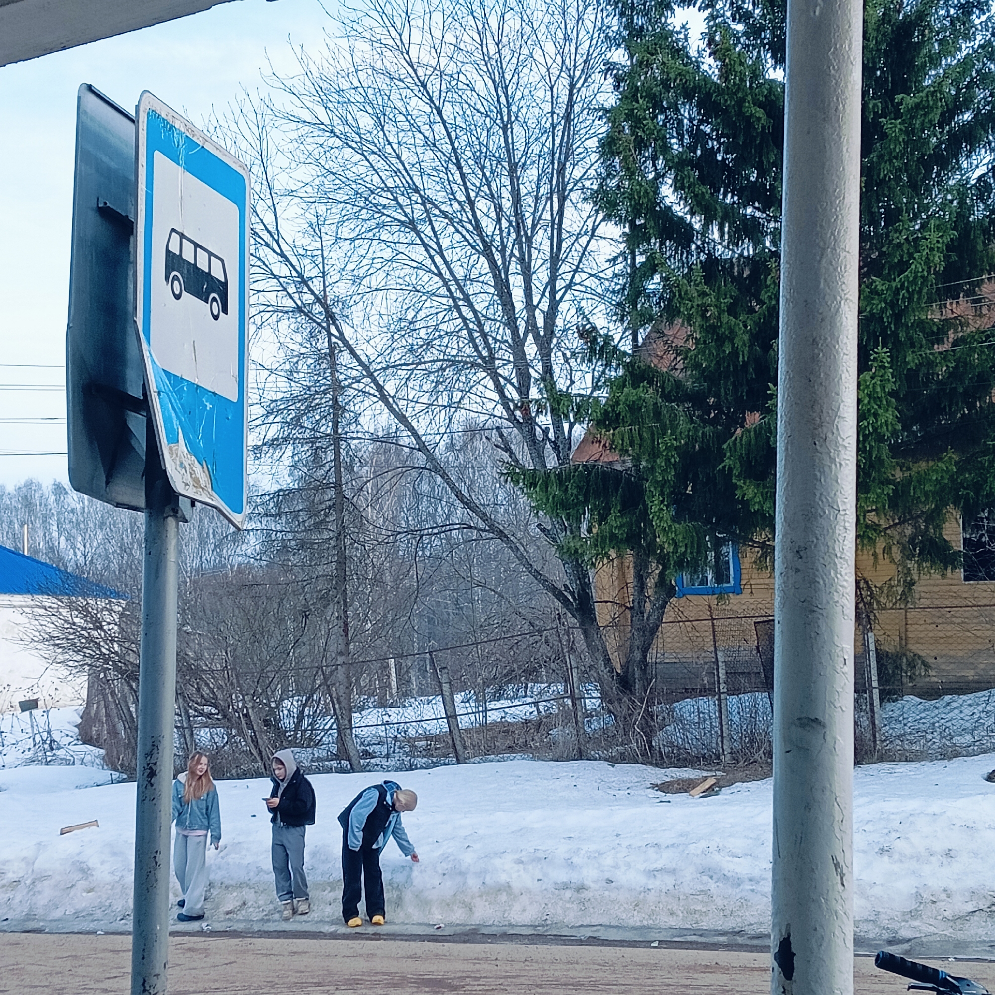 The image shows a bus stop sign in the city of Зипуново, Russia. The sign is blue and white and depicts a bus. It is mounted on a metal pole. The sign is old and damaged. Three people are standing near the bus stop in the snow. One woman is wearing a blue denim jacket, the other a dark hooded jacket and the third is bending over. Behind them is a snow-covered ground, a fence, bare trees, and a wooden building. A metal pole is in the foreground to the right.