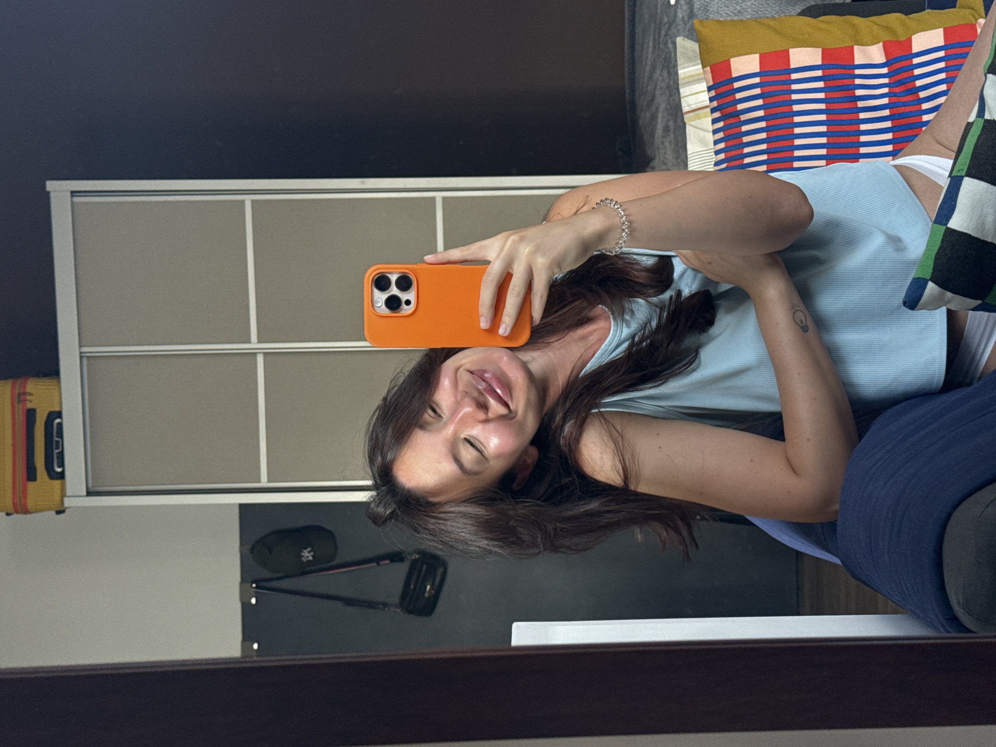 A young woman with long, dark brown hair is taking a selfie in her bedroom. She is wearing a light blue, ribbed tank top and white underwear. Her eyes are closed and she is smiling, tilting her head to the side. She is holding an orange smartphone in her right hand, which is angled to capture her reflection in a large mirror on the left side of the frame. Her left arm is resting on her chest, and she is wearing a silver bracelet on her wrist. A tattoo of a lightbulb is visible on her left forearm.
The background of the image shows a large wardrobe with beige sliding doors. On top of the wardrobe, a yellow suitcase with black accents is visible. To the left of the wardrobe, a dark grey wall features a black baseball cap hanging on a hook, along with a black purse with a gold chain strap. The room appears to be a bedroom, with a bed and pillows visible on the right side of the frame. The overall lighting suggests it is daytime. The specific location in Tel Aviv, Israel, cannot be determined from the visual information provided.