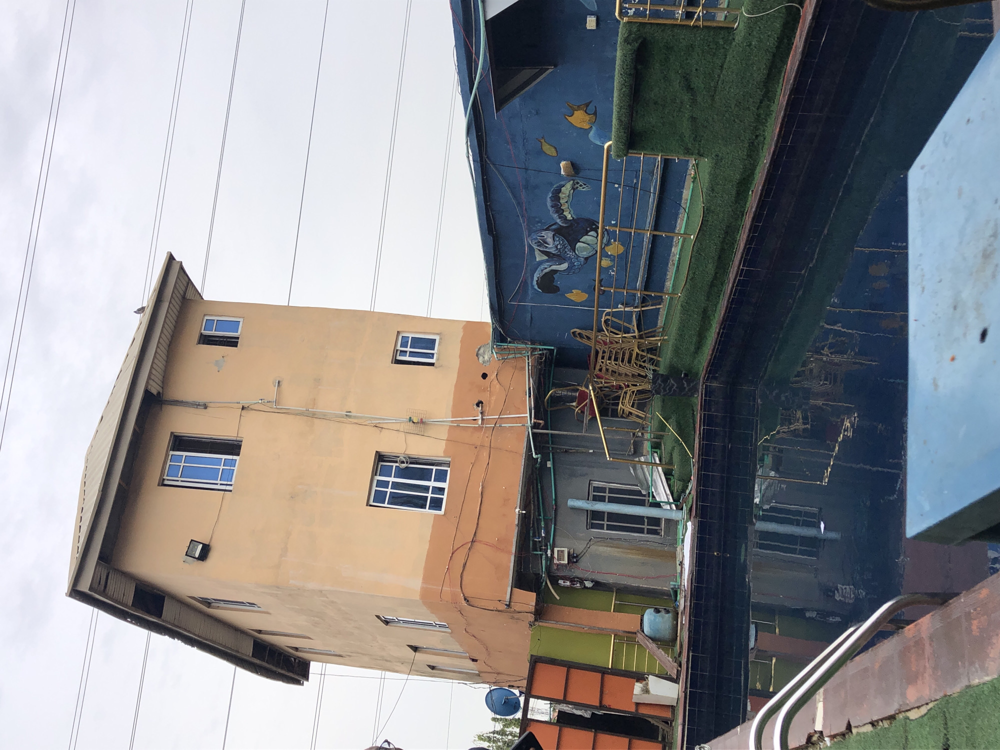 The image shows a tall, peach-colored building in Ado, Nigeria, with multiple windows and exterior lighting. To the right of the building is a structure painted blue with marine life depictions and a patch of artificial grass. Reflections of the surrounding structures are visible in what appears to be a pool or water feature. Power lines are visible against a grey sky. The orientation of the image suggests it might have been rotated. Details in the surrounding area suggest a mixed-use residential and commercial environment.