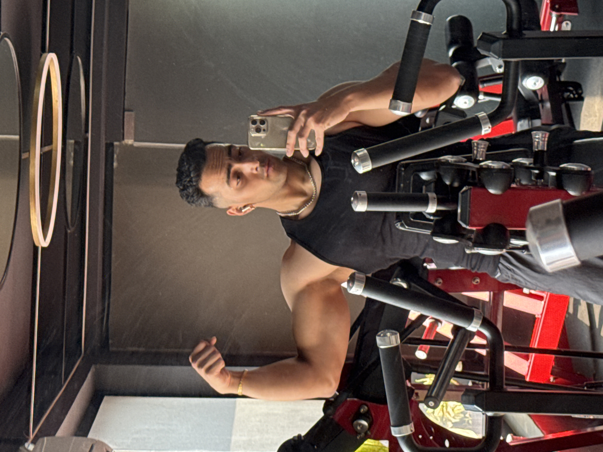 A fit young man is captured in a mirror selfie, flexing his right bicep while holding a silver-colored iPhone. He wears a black sleeveless top, a silver chain around his neck, a gold bracelet on his right wrist, and an earbud in his left ear. His expression is focused and confident.
The activity taking place is a mirror selfie, likely taken in a gym or fitness studio in Kolkata, India. The man is showcasing his physique, specifically his bicep, suggesting a post-workout moment or a demonstration of strength.
The setting appears to be indoors, characterized by professional gym equipment with prominent red and black components and chrome accents, indicating a dedicated fitness environment. To the left of the man's reflection, part of a wall with decorative gold and white rings is visible. The lighting suggests it is daytime. The image itself is rotated 90 degrees clockwise, making the man and background appear on their side.
There is no visible text in the image.