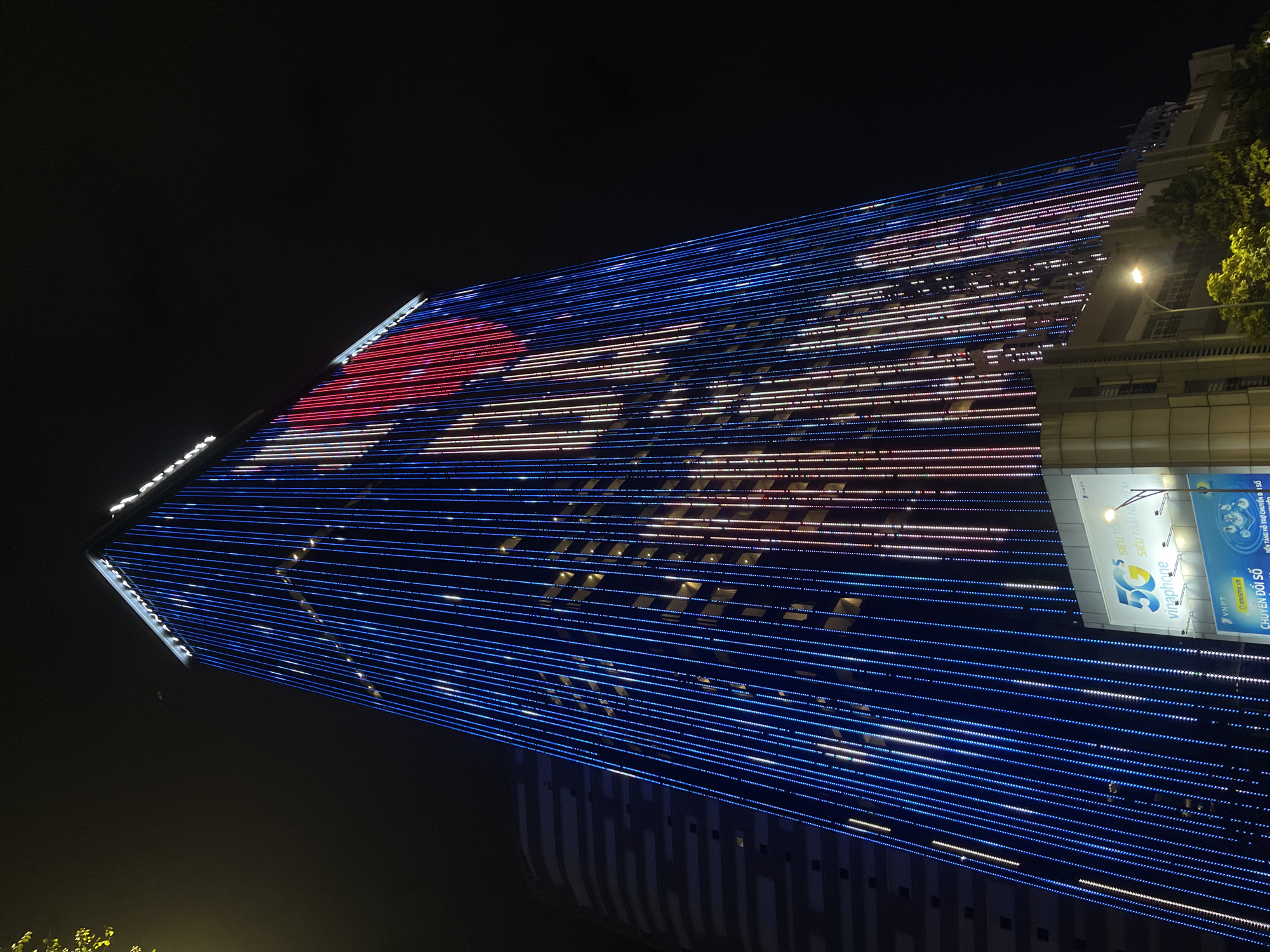 The image captures a nighttime view of the Đà Nẵng Landmark 81, a prominent skyscraper in Đà Nẵng, Vietnam. The building is illuminated with vertical lines of blue and red LED lights, creating a vibrant visual display against the dark sky. Some sections of the building's facade show windows with warm interior lighting, suggesting occupancy. At the base of the building, a large billboard displays the "5G Vinaphone" logo, indicating telecommunications services. The surrounding environment is dark, with hints of other buildings and foliage visible in the background. The perspective is from a low angle, looking up at the towering structure.
