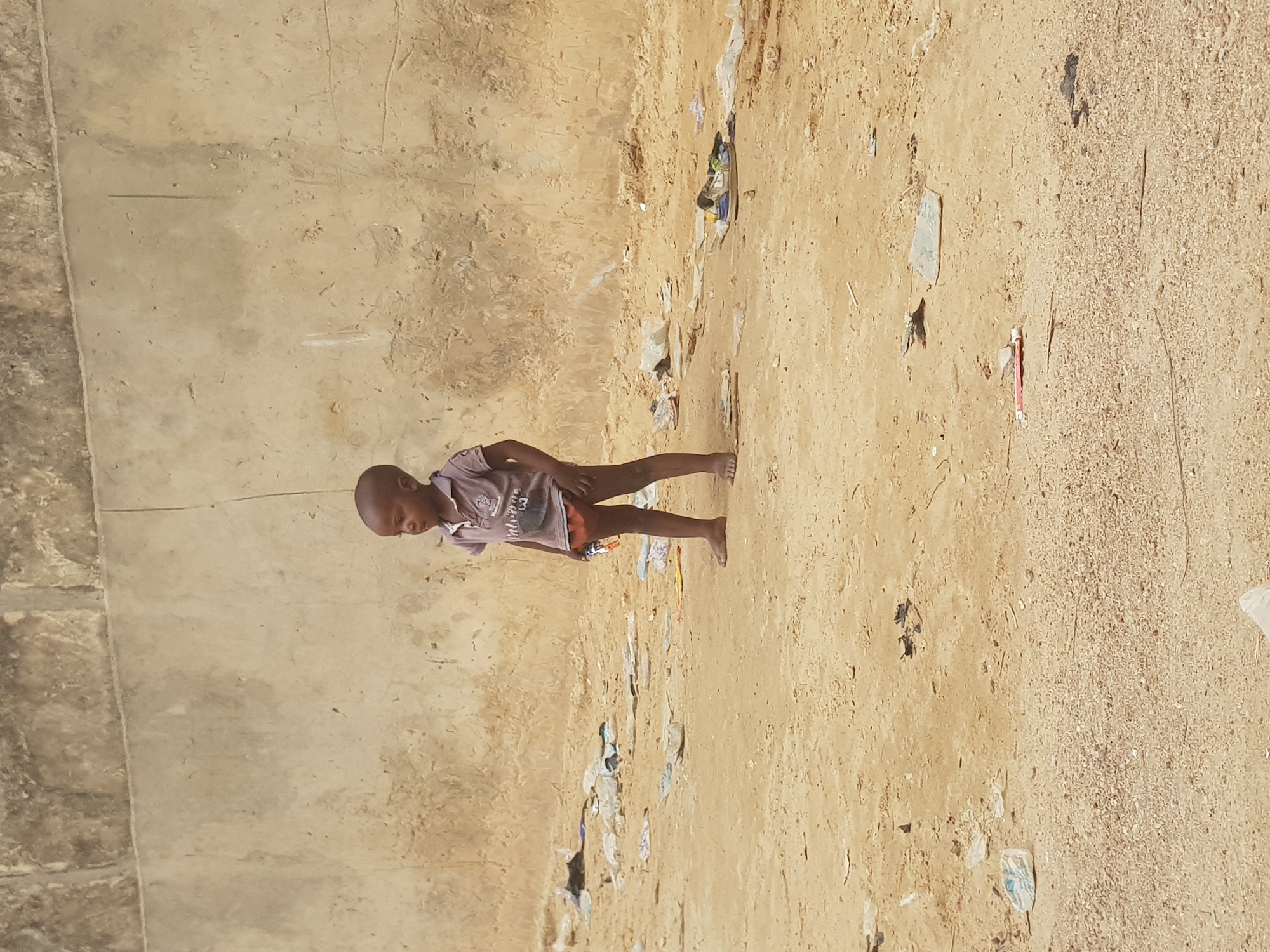 A young, barefoot boy stands on a dirt and sandy surface with some debris near a concrete wall. He wears a collared shirt and short pants. The location appears to be in Dutsinma B, Nigeria.