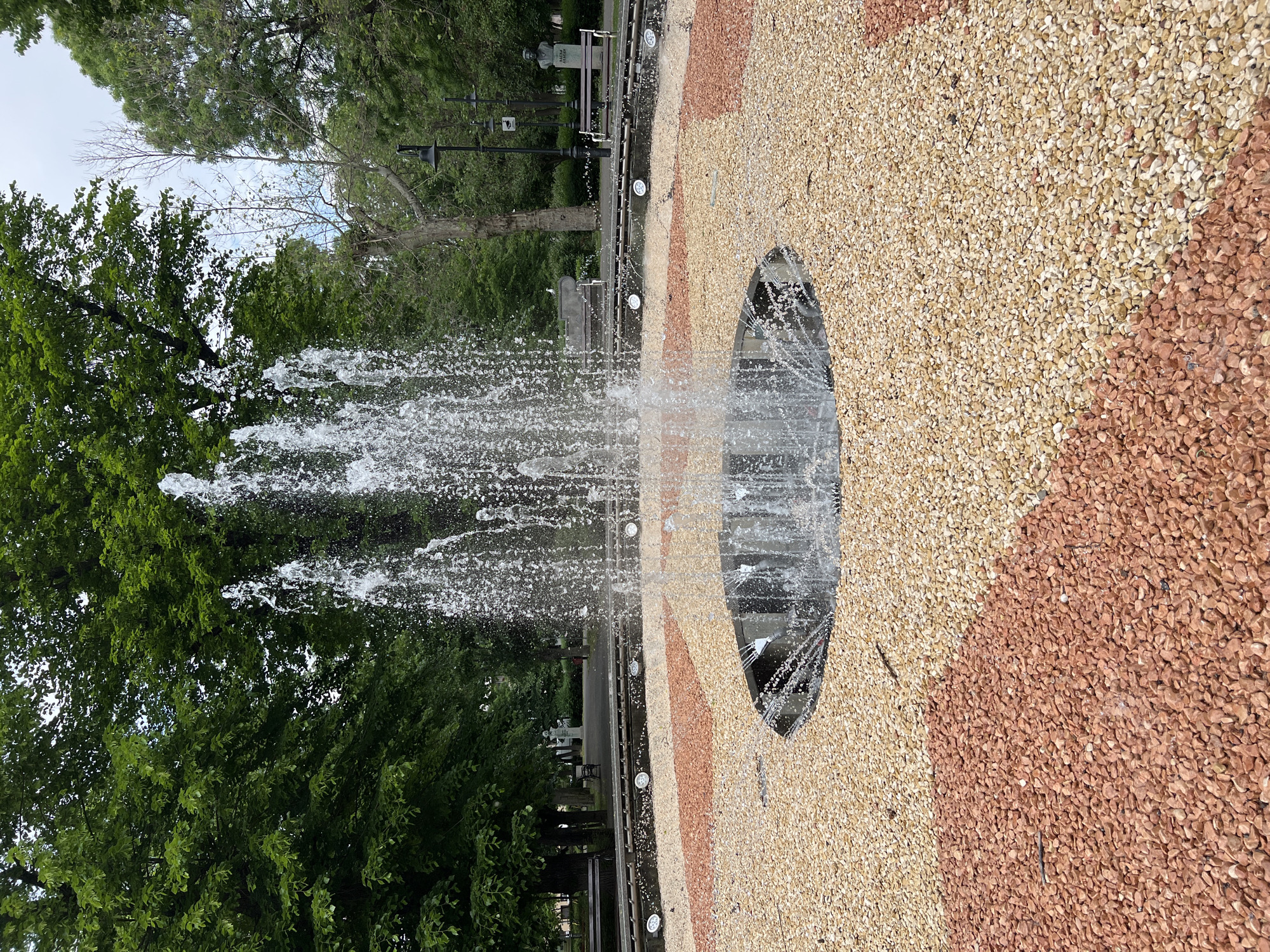 The image captures a fountain in a park setting, likely located in Burgas, Bulgaria. The fountain features jets of water spraying upwards, creating a dynamic display. The surrounding ground is covered with small, multicolored stones arranged in stripes of beige and reddish-brown, creating a decorative pattern. The fountain itself is circular, with a visible black edge. In the background, there are trees with green foliage, benches, and lampposts, indicating a public park area. The photograph is taken from a ground-level perspective, looking upwards at the water jets.