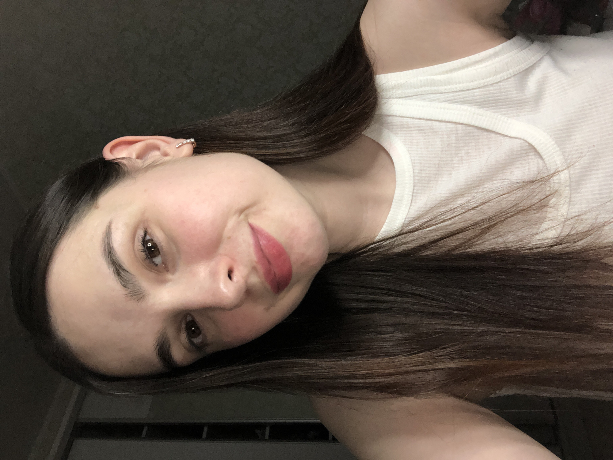 A woman with long, dark brown hair and fair skin is posing for a selfie, tilting her head to the left. She has brown eyes, pronounced eyebrows, and is wearing lipstick. An earring is visible in her left ear. She is wearing a white tank top. The background appears to be a dark, possibly indoor, space. There is no indication of the location being Noviy, Russia, within the image.