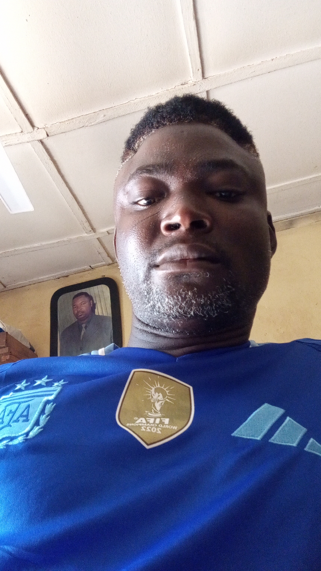 A dark-skinned man is captured in a close-up selfie, looking directly at the camera with a neutral expression. He has short, dark, textured hair with lighter highlights around the hairline, and a salt-and-pepper beard. His forehead appears slightly sweaty.
He is wearing a blue short-sleeved soccer jersey. On the left chest, a light blue crest with the letters "AFA" and three stars is partially visible. On the right chest, there is a gold hexagonal patch with the FIFA World Champions 2022 logo, featuring the mirrored text "FIFA WORLD CHAMPIONS 2022". Light blue Adidas stripes are visible on the right shoulder of the jersey.
The man is indoors, likely in Sabon Tasha, Nigeria. The background features a light-colored ceiling with visible planks or beams, and a light yellow wall. To the left side of the frame, a framed picture hangs on the wall, depicting another dark-skinned man with short hair, wearing a suit and tie, looking forward. The overall lighting suggests it is daytime.