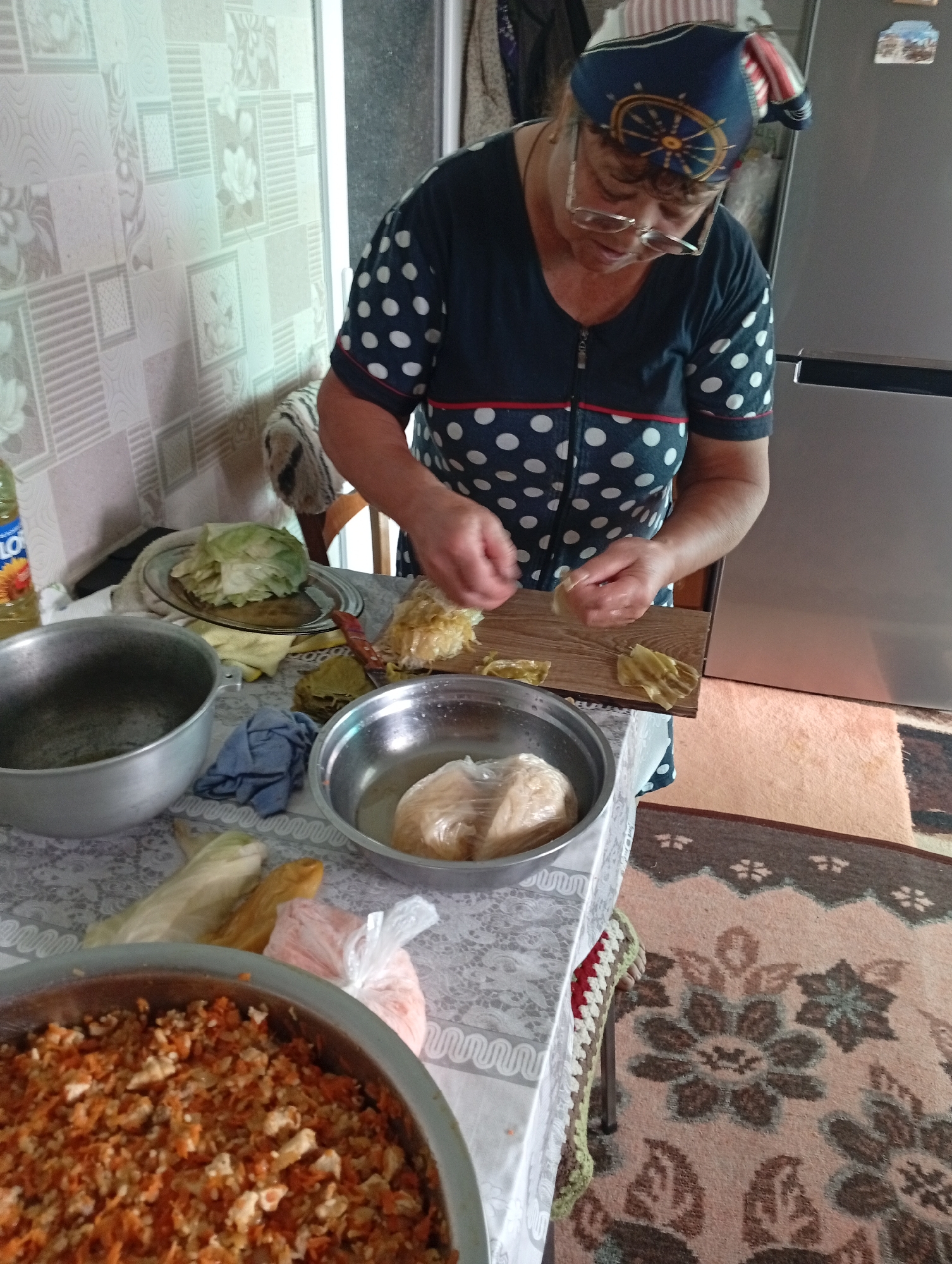 In Pănășești, Moldova, a woman is preparing food in a home kitchen. She is standing at a table, wearing a dark blue and white polka dot shirt and a patterned headscarf. Her glasses are perched on her nose as she focuses on her task. On the table, there are several items: a large metal pot, a metal bowl filled with a light-colored filling wrapped in plastic, a plate with cooked cabbage leaves, and a wooden cutting board with more cabbage leaves and what appears to be a filling. A bottle of sunflower oil is visible on the left side of the image, and a large bowl of chopped carrots and other ingredients is in the foreground. The background shows a wall with patterned wallpaper and a refrigerator. The scene suggests the preparation of stuffed cabbage rolls, a common dish.
