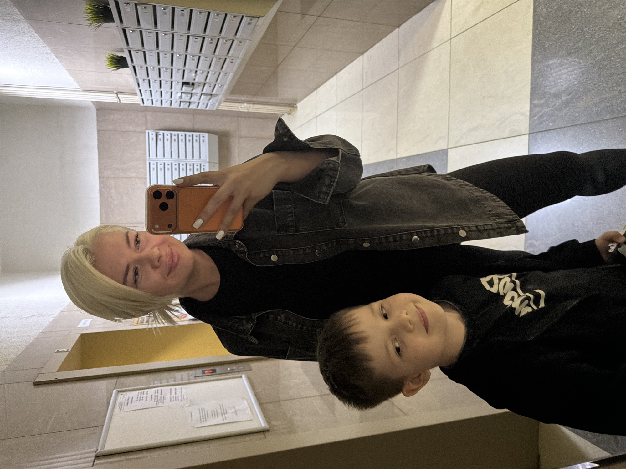 The image depicts a woman and a young boy in what appears to be the lobby of an apartment building. The woman, with blonde hair, holds an orange-cased smartphone, presumably taking a selfie. She wears a black top and a dark denim jacket. The boy, sporting short brown hair and a black sweatshirt with white lettering, stands next to her. Behind them, the environment includes an elevator door with a call button, a wall-mounted notice board, rows of mailboxes, and tiled flooring. The city and specific location within that city are not discernible from the image.