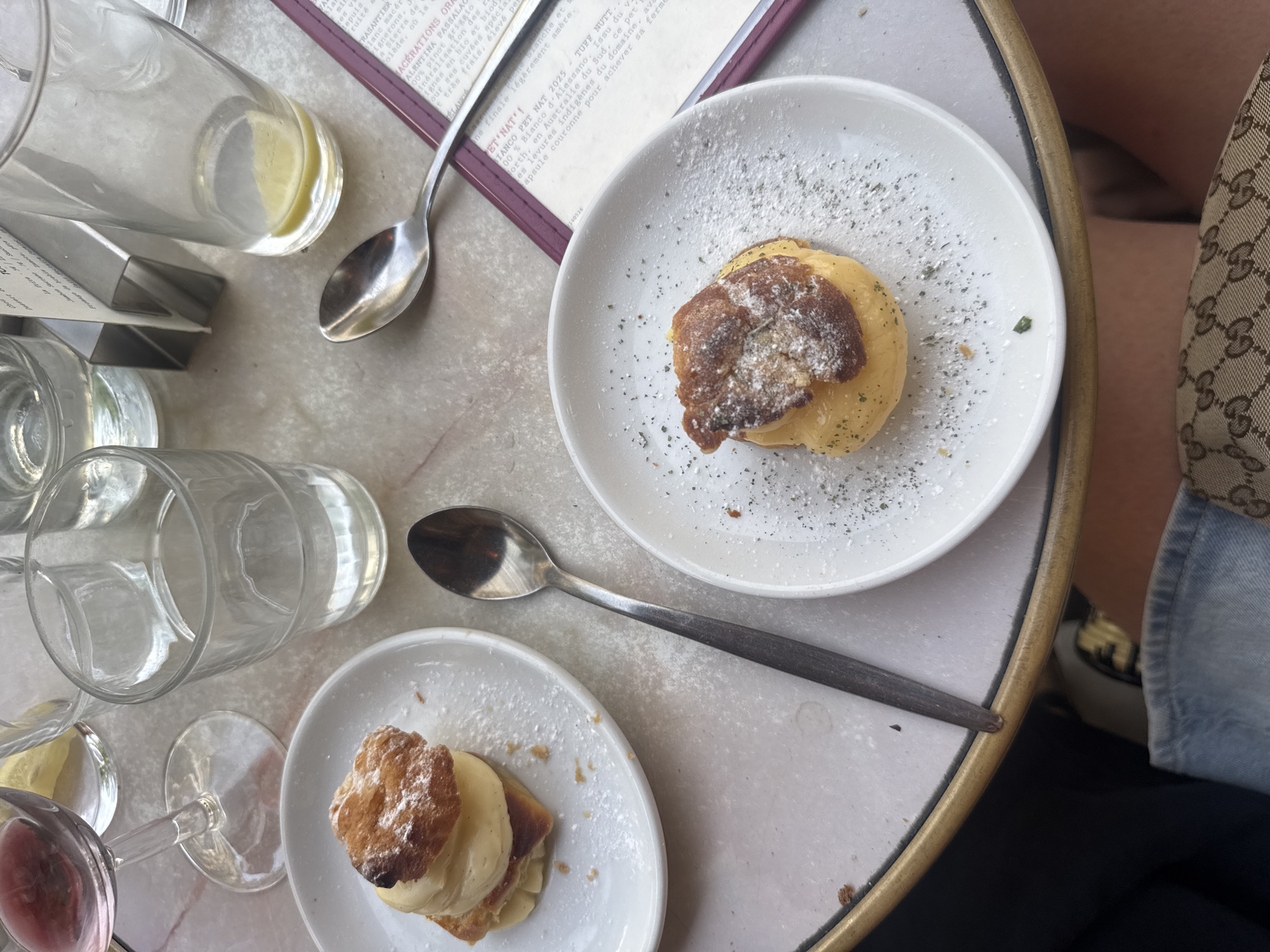 This overhead shot captures a delightful scene on a marble-topped table, likely in a Parisian cafe, given the presence of a menu with French text. Two small, elegantly presented desserts, sprinkled with powdered sugar and green herbs, sit on white plates. One dessert is topped with a dark, flaky pastry, while the other has a lighter, golden-brown pastry. Silver spoons rest near each plate, suggesting a leisurely meal.
Surrounding the desserts are several glasses, some containing water with a slice of lemon, and a stemmed glass holding a pinkish liquid, possibly wine. The table itself is slightly textured, with a golden rim. In the background, a person's legs, clad in denim and a distinctively patterned designer fabric, suggest a relaxed and possibly upscale setting. The menu, partially visible, contains wine descriptions, indicating a focus on beverages. The lighting suggests daytime, possibly late afternoon. The overall impression is one of quiet enjoyment and indulgence in a charming European setting.