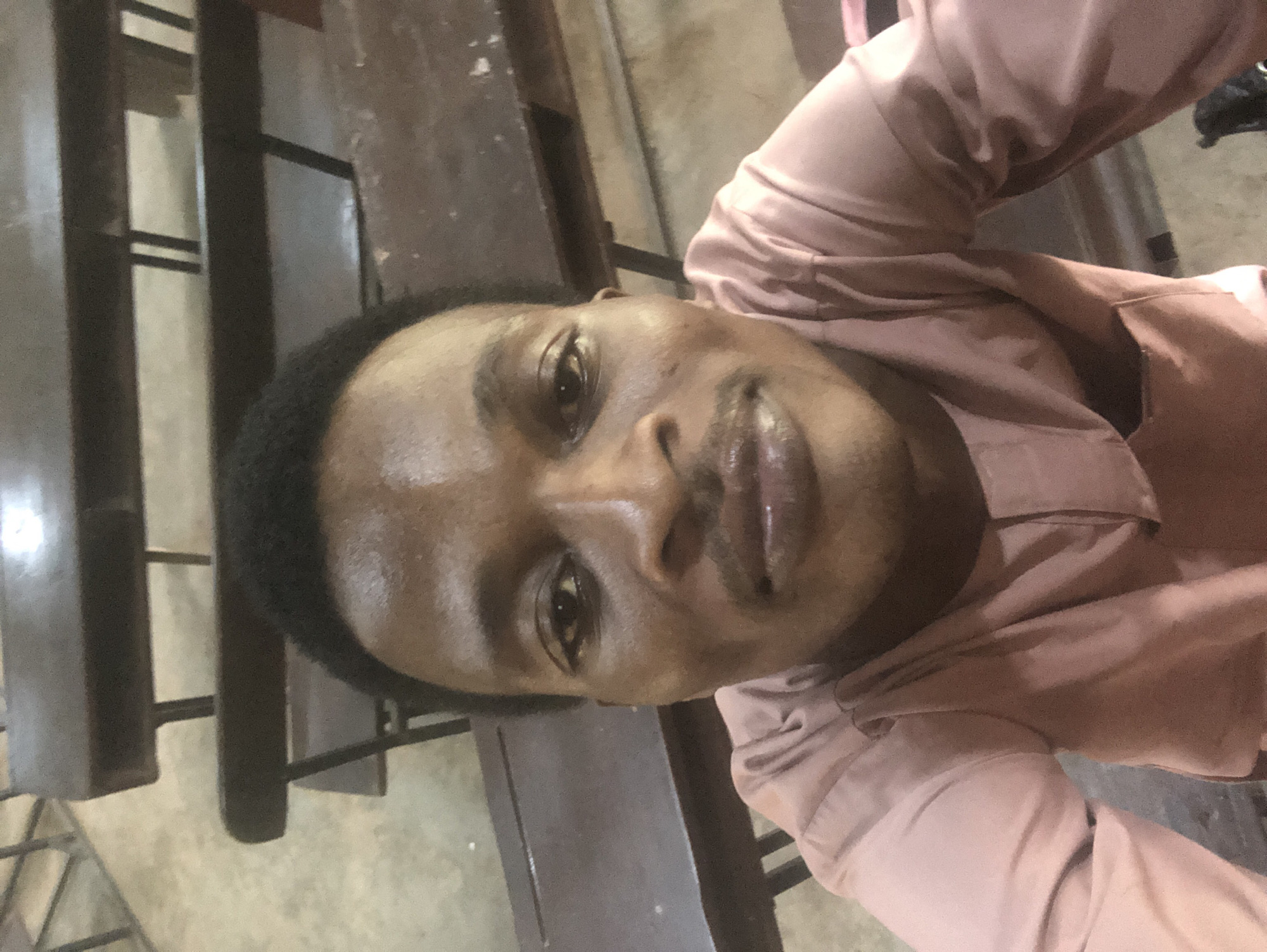 A close-up selfie of a young Black man shows him looking directly at the camera. He has dark, short hair, brown eyes, and a thin mustache. He is wearing a pale pink, long-sleeved shirt with a Mandarin collar. The background consists of dark wooden benches, suggesting he is in a hall or auditorium. The lighting is dim.
