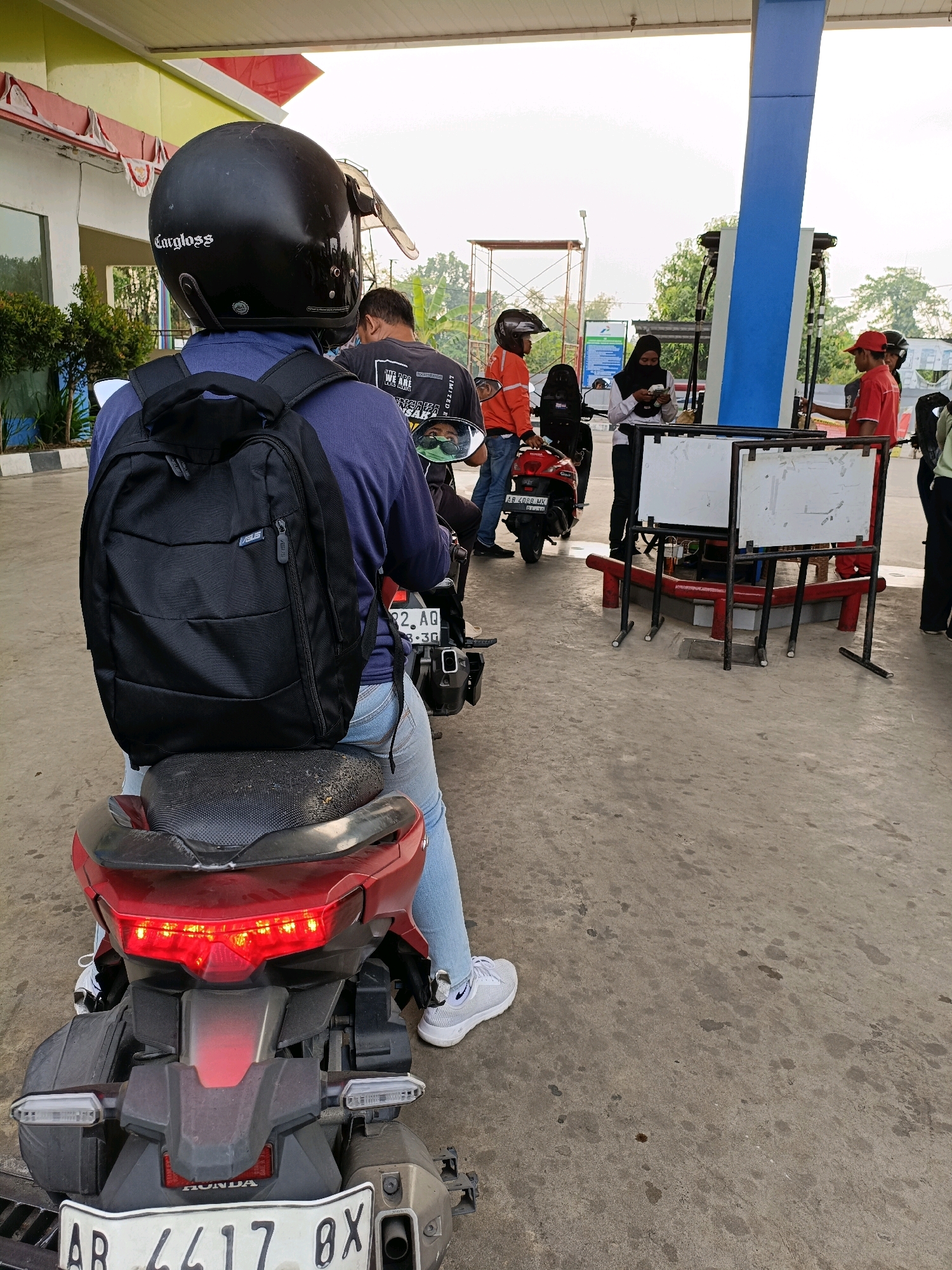 The image depicts a scene at a gas station in Maguwoharjo, Indonesia. A person wearing a black helmet and a blue shirt with a black backpack is seated on a red motorcycle, facing away from the camera. In the background, several other individuals are present. One person in an orange jacket stands near a red motorcycle. Another person wearing a hijab is holding money and looking at their phone. A person in a red shirt, likely a gas station attendant, is interacting with a customer near a blue and white gas pump. The ground is concrete, with some oil stains visible. Trees and a building are present in the background. The overall atmosphere suggests a typical day at a busy gas station.