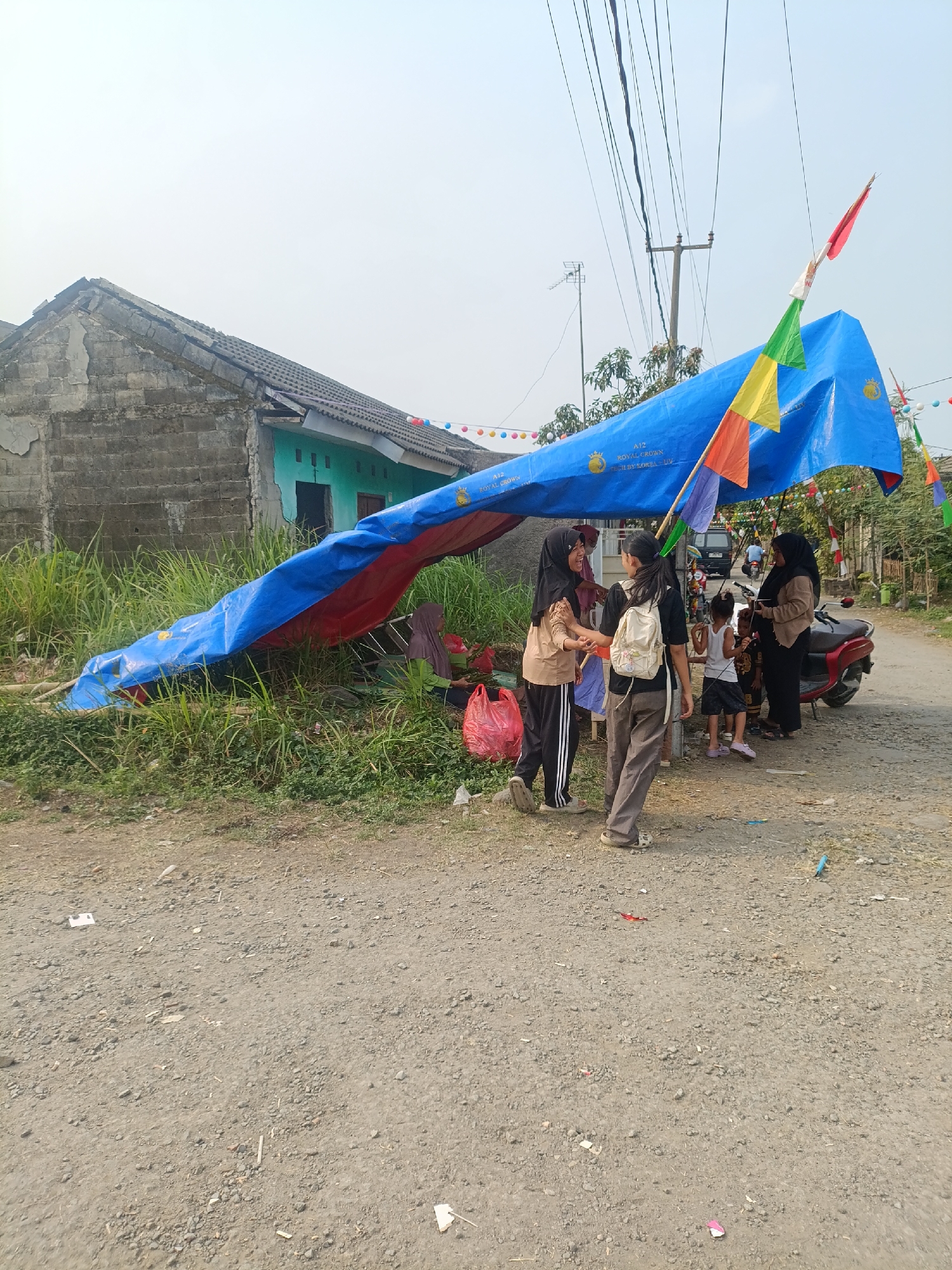 In Cariu, Indonesia, a temporary shelter is set up using a large blue and red tarp, supported by what appears to be a metal frame or poles. The shelter is located outdoors, on a gravel and dirt path adjacent to overgrown grass and a damaged, gray cinder block structure with a green-painted house in the background. Several people, including adults and children, are gathered under or around the shelter. One adult woman wearing a hijab is interacting with a child. Another woman, also wearing a hijab, is standing next to a red motorcycle. Two younger women are facing each other, one of whom is wearing a backpack. The scene is decorated with small, colorful flags and banners, suggesting a festive occasion or event. Overhead, power lines and a utility pole are visible against a hazy sky.