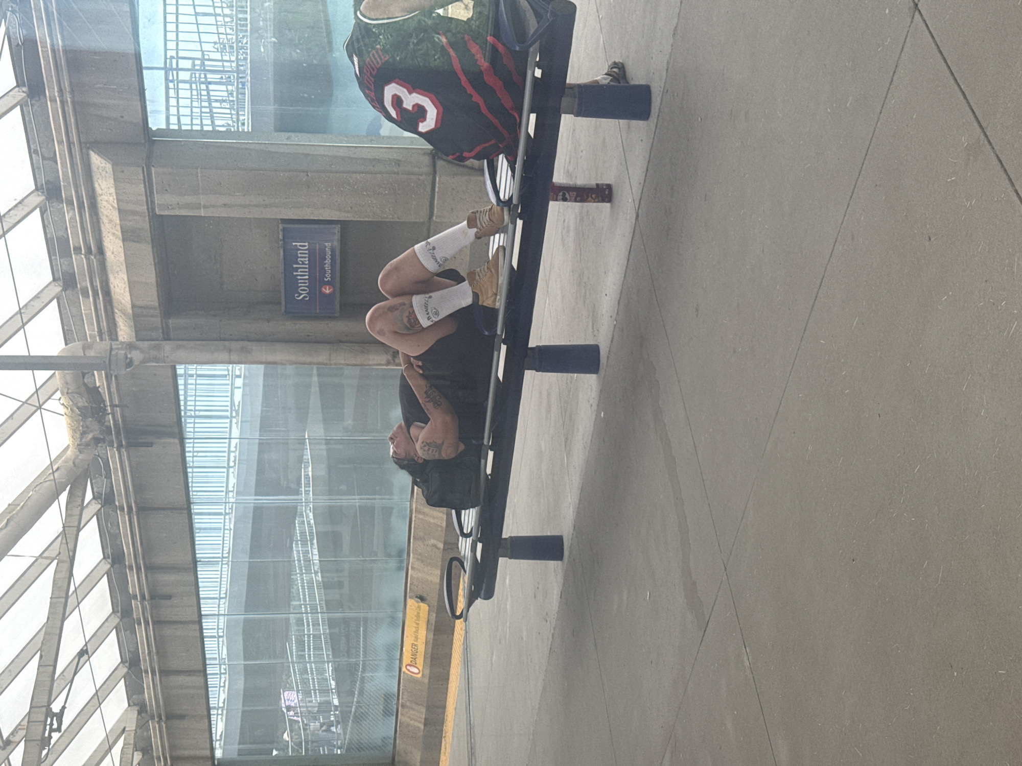 A person is lying on a bench at the Southland LRT station in Calgary, Canada. The person is wearing shorts, a dark t-shirt, white socks, and tan shoes, with visible tattoos on their arms and legs. A black backpack is on the bench next to the person. To the right, another person is seated on the bench, wearing a black and red sports jersey with the number "3" on the back. Behind the bench, a sign indicates "Southland" and "Southbound." The station features a covered waiting area with a translucent roof and large glass windows. A yellow "DANGER" sign is visible near the edge of the platform.