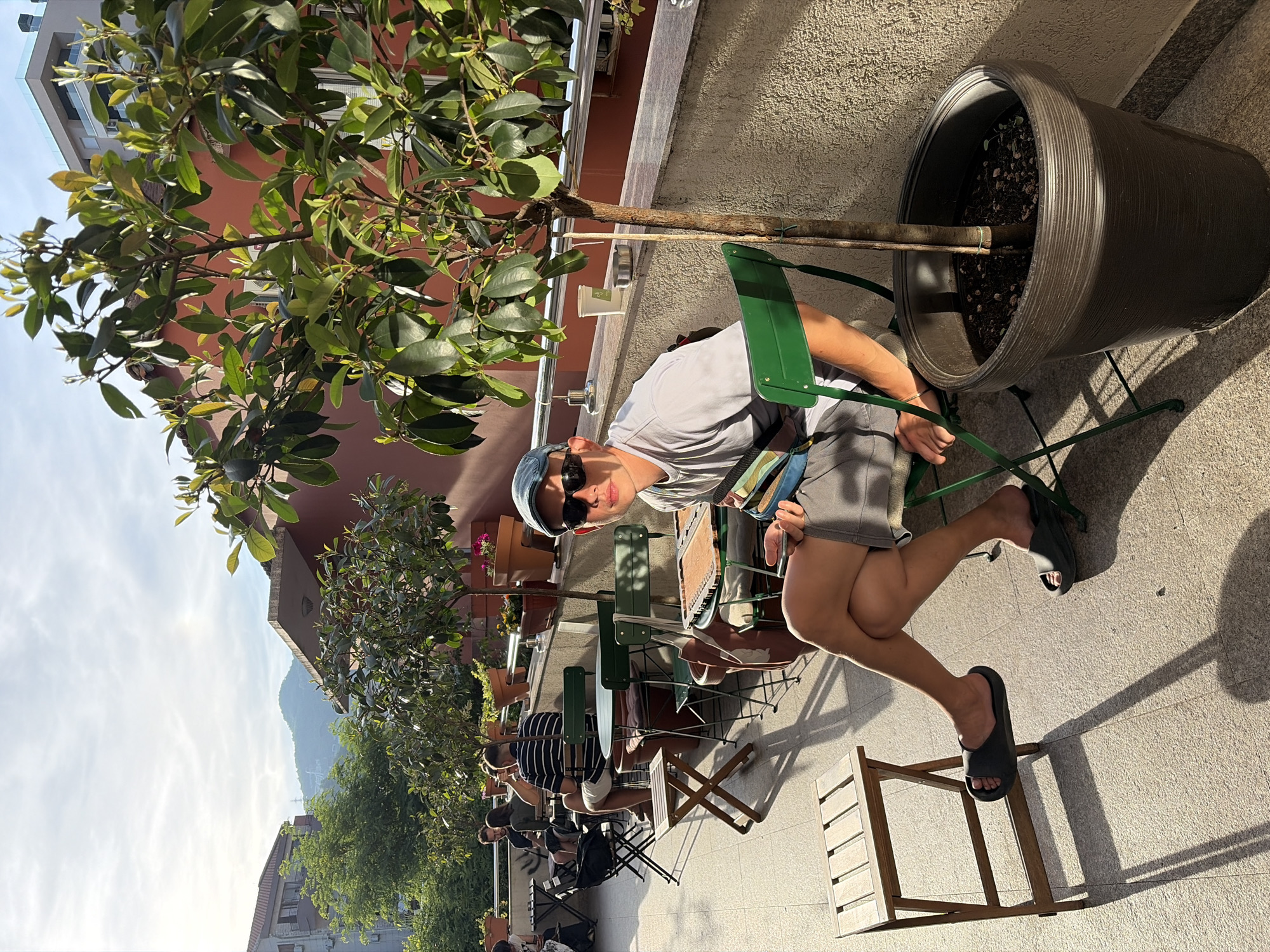 The image captures a vibrant outdoor cafe scene in Budva, Montenegro, on a sunny day. The perspective is slightly rotated, showing a man prominently in the foreground, seated on a dark green metal chair. He wears a light gray t-shirt, gray shorts, dark flip-flops, a blue baseball cap, and dark sunglasses, looking directly towards the viewer with a neutral expression. His legs are crossed, and one hand rests on his knee.
To his immediate right, a tall, leafy tree is planted in a large, dark gray pot, its trunk supported by a stake. The ground is paved with light-colored tiles, exhibiting long, distinct shadows from the chairs, tables, and the potted plant, indicating bright sunlight, likely late morning or afternoon. Near the man's feet is a small, light-colored wooden stool.
In the mid-ground, several other individuals are seated at similar green metal tables and wooden folding tables, also enjoying the outdoor setting. They are casually dressed. A low wall or balcony runs behind them, adorned with more potted plants.
The background reveals a picturesque setting with buildings featuring reddish-brown walls and tiled roofs, characteristic of the Mediterranean region. Lush green trees and foliage are scattered throughout the urban landscape. In the far distance, majestic mountains rise under a bright, partly cloudy sky, with a visible sun glare. The overall atmosphere suggests a relaxed, leisurely day at an outdoor establishment. No legible text is visible in the image.