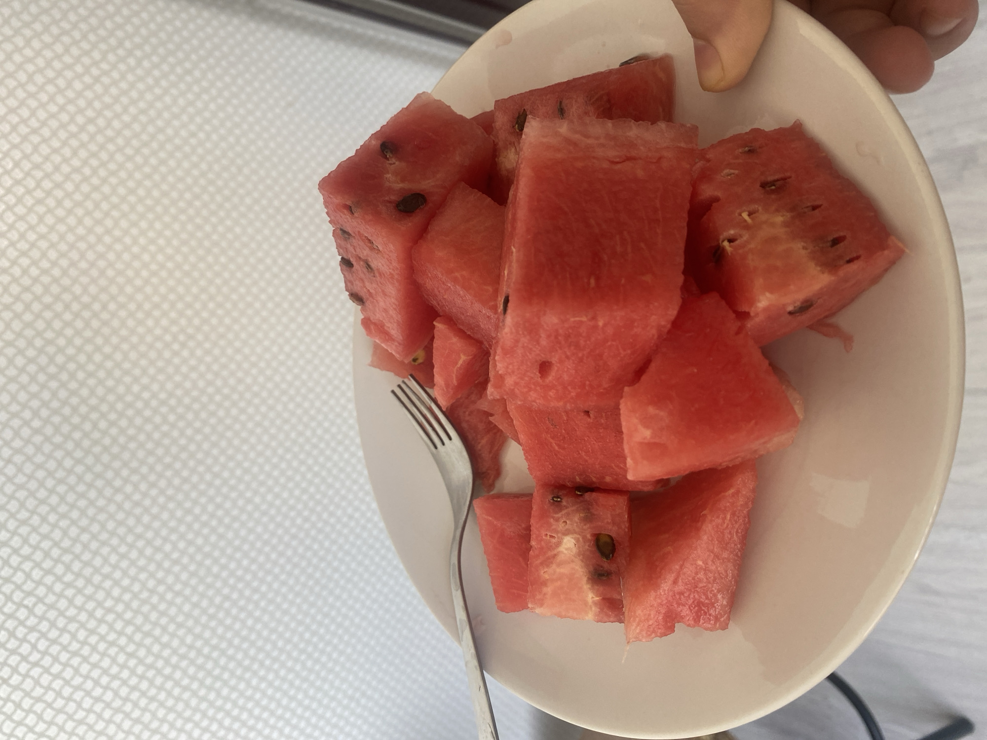 A white plate filled with cubed watermelon is positioned on a white surface. A metal fork rests on the plate, its tines lightly touching some of the watermelon pieces. A hand is visible on the right side of the frame, holding the plate. The background is a light-colored wall with a subtle textured pattern. The scene appears to be indoors, potentially in a domestic setting or a casual dining area. The image does not provide specific clues to determine its location within Chișinău, Moldova.