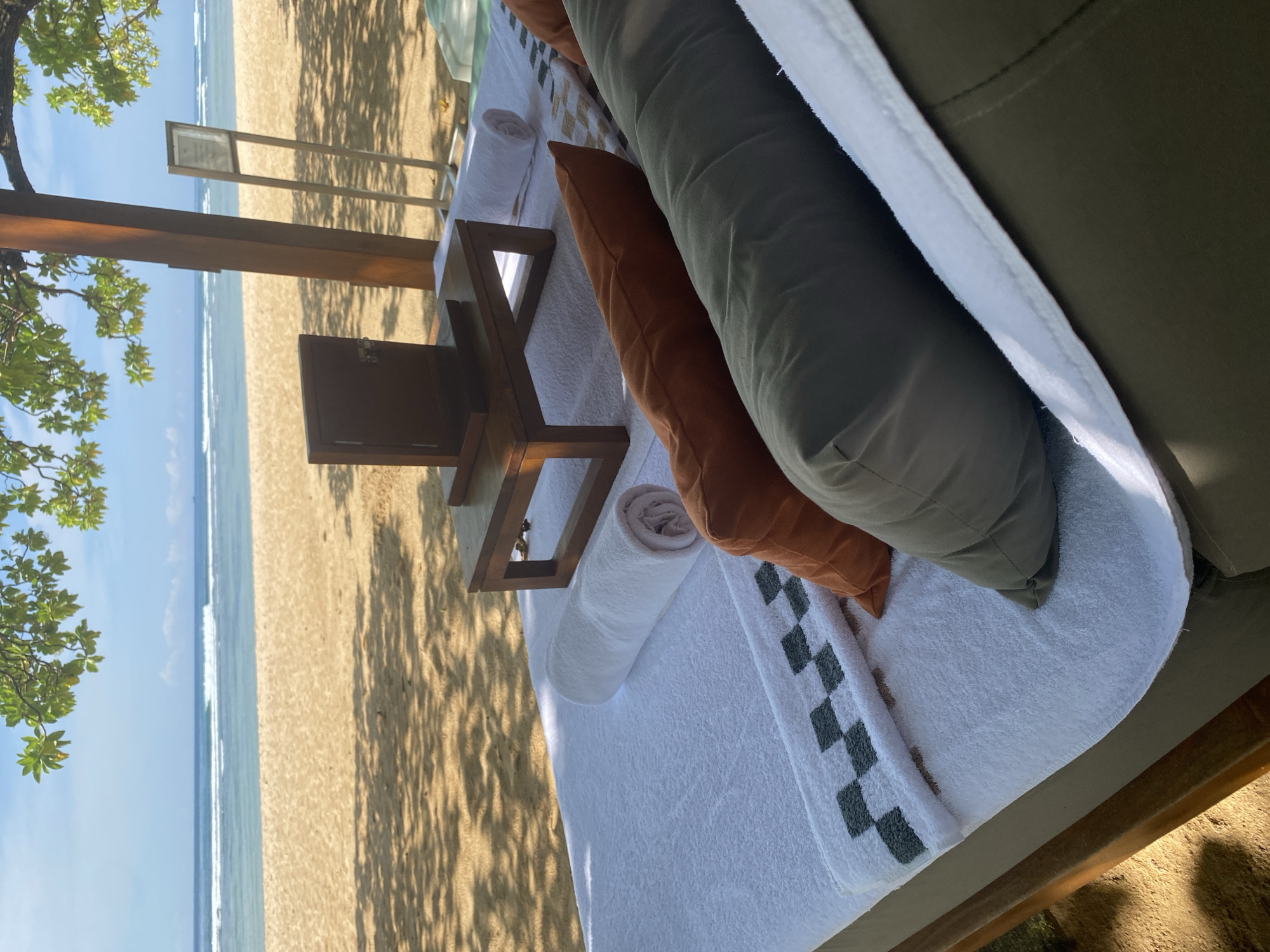 The photograph shows a beach setting, likely taken from a resting area. The scene features a sandy beach with gentle waves meeting the shoreline. In the foreground, there is a lounger with white linens, decorative pillows in brown and gray, and rolled-up towels. A small wooden table is positioned next to the lounger. The composition suggests a tranquil, vacation-oriented atmosphere. Given the location of Nusa Dua, Indonesia, this scene is likely at a resort or private beach. There are no people visible in the shot. The scene conveys relaxation and beachside leisure.