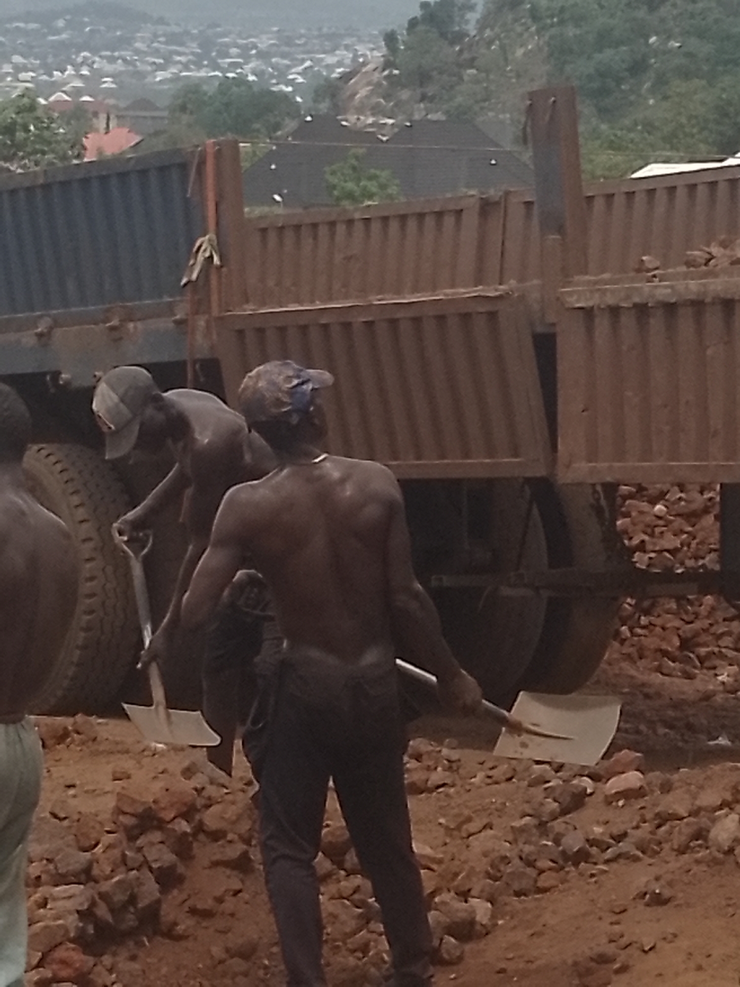 The image captures a scene of manual labor at what appears to be a construction or excavation site. In the foreground, at least three men, bare-chested and seemingly engaged in strenuous work, are visible. Two of the men are prominently holding large shovels, actively moving reddish-brown dirt and rocks from the uneven ground. One man on the left is partially cropped but also appears to be involved in the work. One worker wears a baseball cap, and another has a blue head covering.
Behind the men are two large, heavy-duty dump trucks with tall, ribbed metal sides. The trucks appear to be either loading or unloading material, and their large tires are partially visible. The ground is covered in loose earth, gravel, and chunks of rock.
In the mid-ground and background, a sprawling settlement of numerous buildings, possibly houses or other structures, spreads across undulating hills under a hazy sky. The distant landscape suggests a populated, possibly urban or semi-urban, area.
The time of day appears to be daytime, though the lighting is soft and diffused, indicating either an overcast sky, early morning, or late afternoon. There are no strong shadows. The atmosphere seems dusty or hazy. No visible text is present in the image. The overall scene depicts hard manual labor in a developing or industrial setting.
