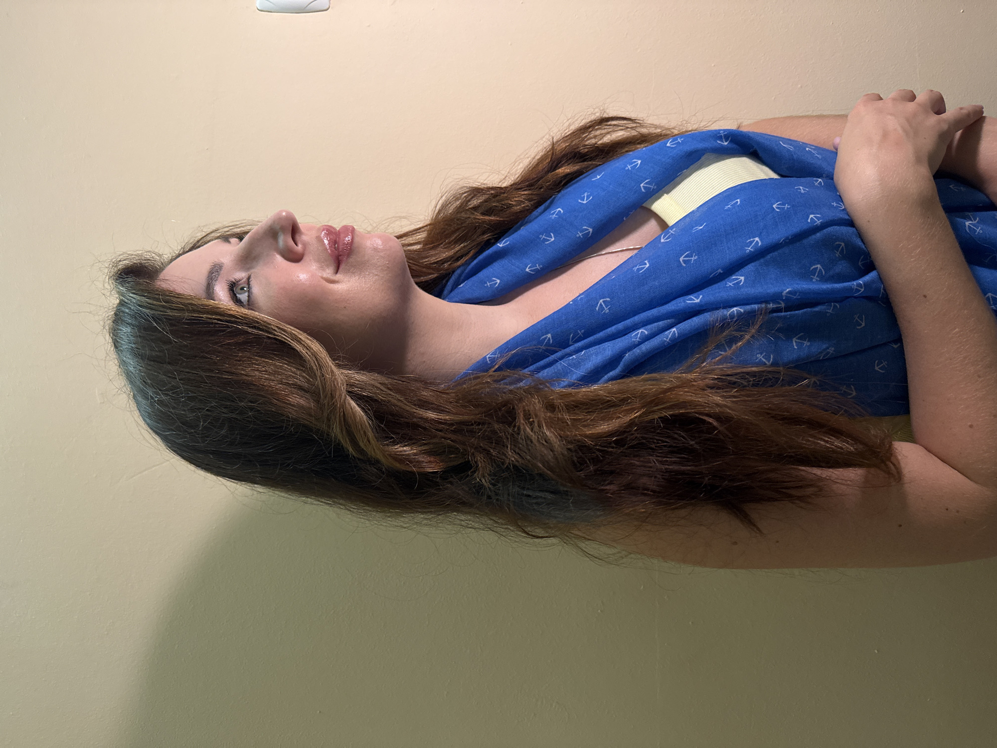 An image shows a woman posing against a pale yellow wall. She is looking up and to her right, with her mouth slightly open and her lips glossy. Her long, wavy brown hair falls over her shoulders and chest. She is wearing a bright blue scarf with a white anchor pattern, layered over a yellow top. Her left arm is bent, with her hand resting on her right arm. A faint shadow is cast on the wall behind her. The image appears to be an indoor portrait. There is no specific information in the image to indicate its location in Odesa, Ukraine.