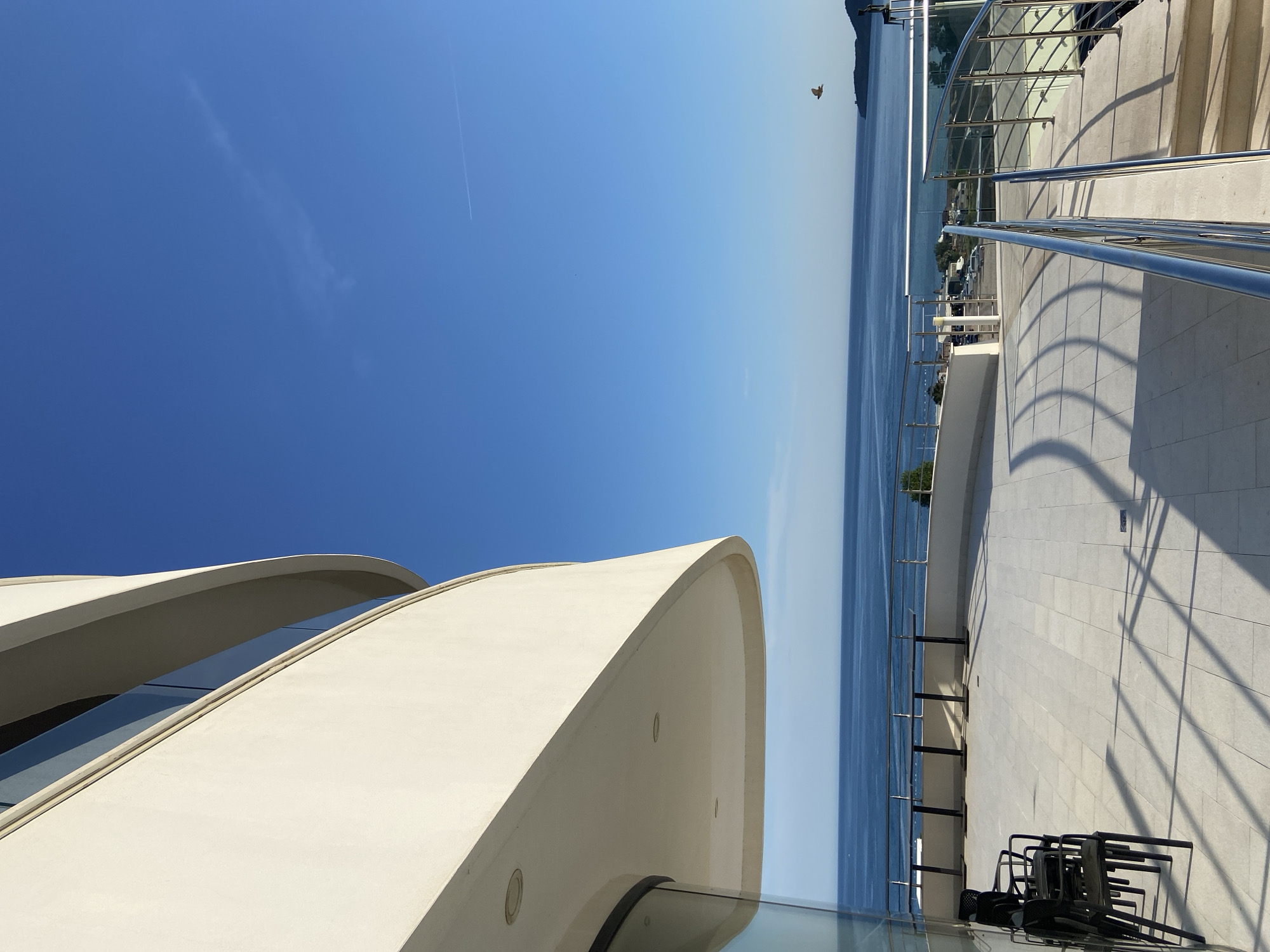 A sunlit, high-angle, outdoor daytime shot of a modern building in Boreti, Montenegro. The building features curved, white concrete elements with glass panes. The Adriatic Sea stretches to the horizon. A stainless steel railing runs along a tiled terrace with stacked chairs. A bird flies above the sea, and a contrail is visible in the sky.