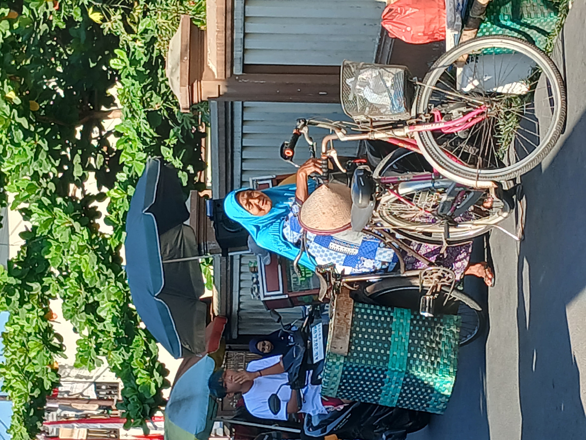 The image captures a street scene in Surakarta, Indonesia, on a sunny day. In the foreground, an elderly woman wearing a blue hijab and a patterned blue and white dress is seated on a bicycle. She has a straw basket on the handlebars. Beside her, a pink bicycle with a wire basket is parked. To the left, a young man in a white t-shirt and red shorts is seated on a motorbike, smoking. Behind him, another person wearing a dark hijab is also on the motorbike. A large woven basket with a green and blue pattern is attached to the back of the motorbike. In the background, there are some stalls with umbrellas, a building with vertical red and white stripes, and lush green foliage from trees. A brown wall and fence are also visible. A red plastic bag is placed near the bicycles on the right side of the image. The ground is paved with asphalt.