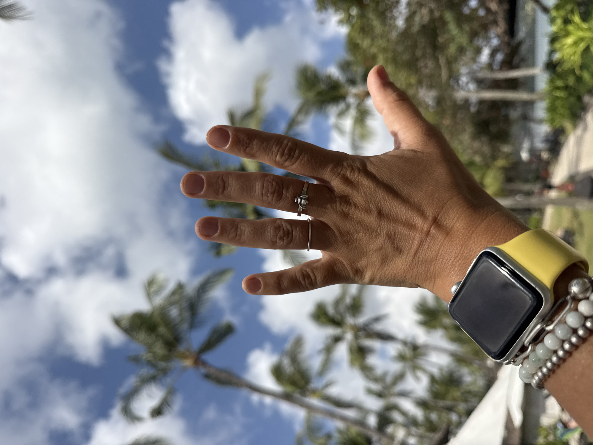 An over-the-shoulder, first-person perspective shot focuses on a person's left hand and wrist against a backdrop of palm trees and a cloudy blue sky, suggesting a tropical location like Kuta Selatan, Indonesia. The hand is open, with fingers extended upwards, and adorned with two silver rings. On the wrist, a smartwatch with a bright yellow band is visible, accompanied by stacked bracelets. The background is blurred, with the fronds of several palm trees dominating the frame, interspersed with patches of blue sky and white clouds. The overall impression is one of leisure and outdoor activity.