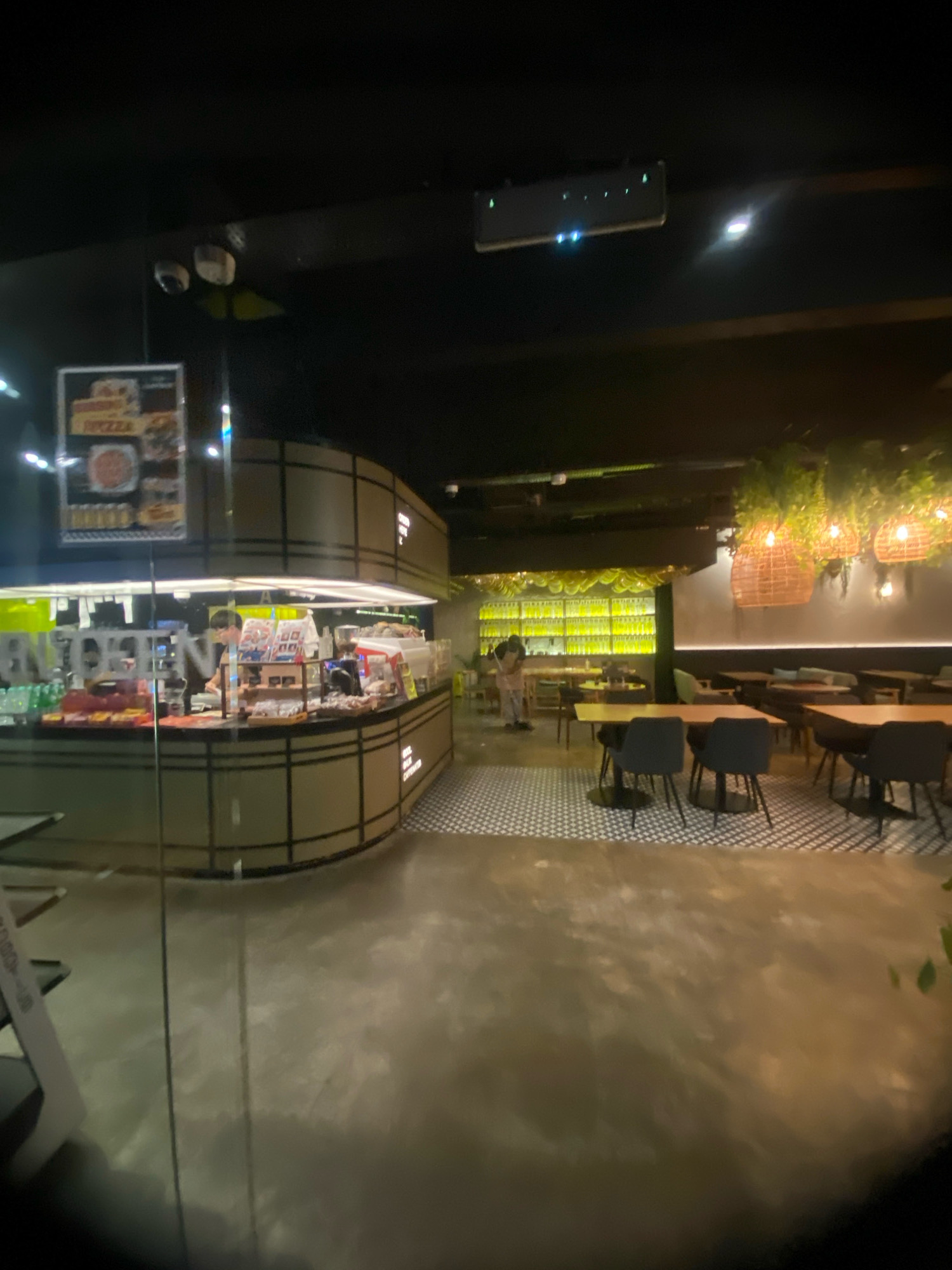 The image depicts the interior of a dimly lit restaurant or cafe, likely located in Kuala Lumpur, Malaysia, given the context. The scene is captured from a low-angle perspective, looking across a polished concrete floor towards a food service counter and seating area.
In the foreground, the reflective concrete floor shows subtle patterns of light and shadow. To the left, a portion of a curved counter is visible, with a menu board displaying pictures of food items like pizza. Behind the counter, a person wearing glasses and a dark shirt is standing and appears to be working, with various food items and containers on display. The counter area is brightly lit, contrasting with the overall dim ambiance.
Further into the space, the seating area is arranged with tables and chairs. Dark-colored chairs with black circular bases are positioned around light-colored tables with similar bases. The floor in this area features a black and white checkered pattern. Beyond the seating area, a wall is lined with shelves holding numerous bottles, illuminated by a bright yellow light. Hanging from the ceiling above the seating area are decorative elements consisting of woven baskets and lush green plants.
Several security cameras are visible mounted on the ceiling. The overall atmosphere suggests a modern, contemporary establishment.